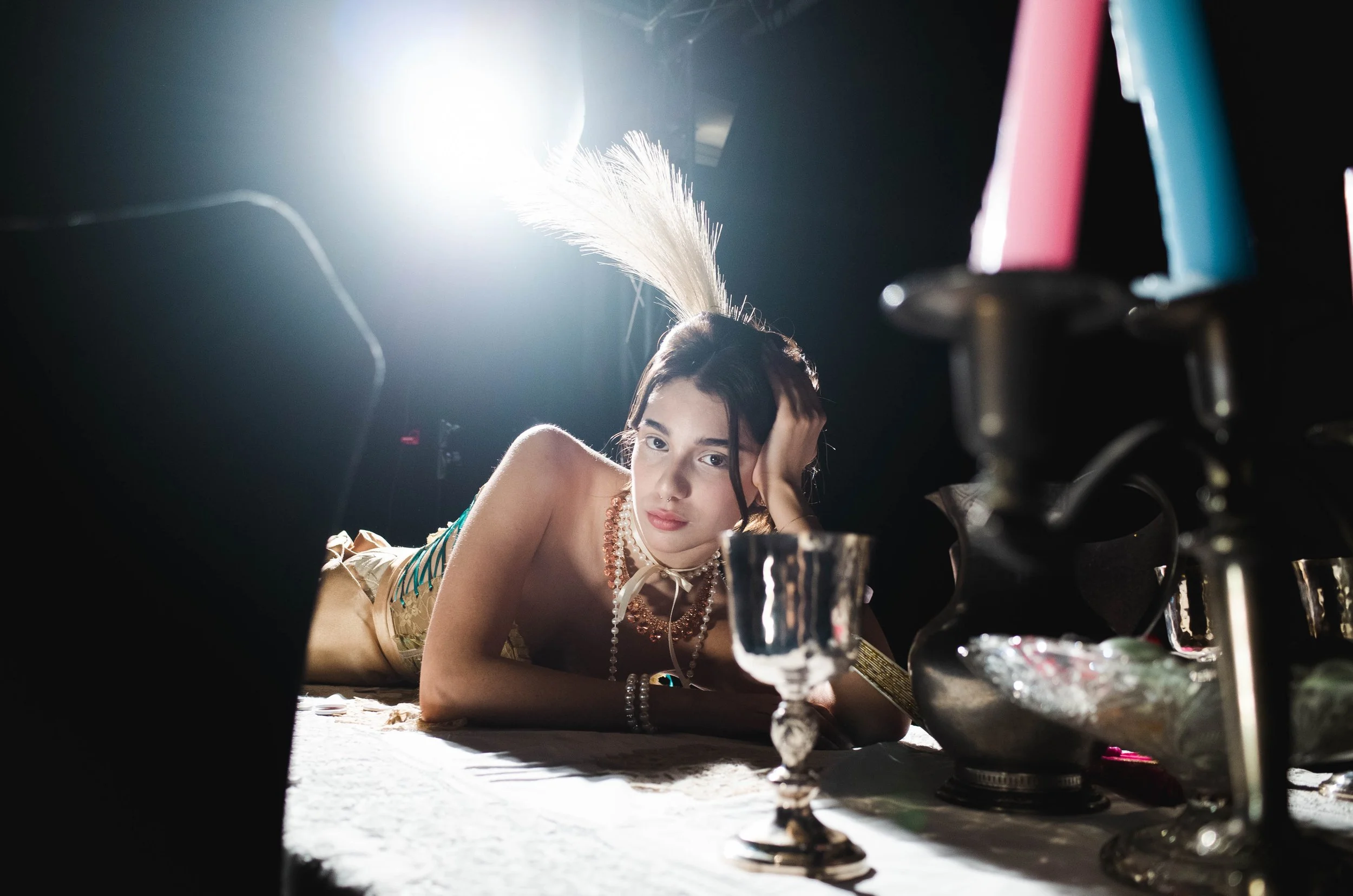 Young woman lying on a table with jewelry and a feather in her hair, surrounded by candle holders and ornate dishes, with a bright light behind her in a dark setting.