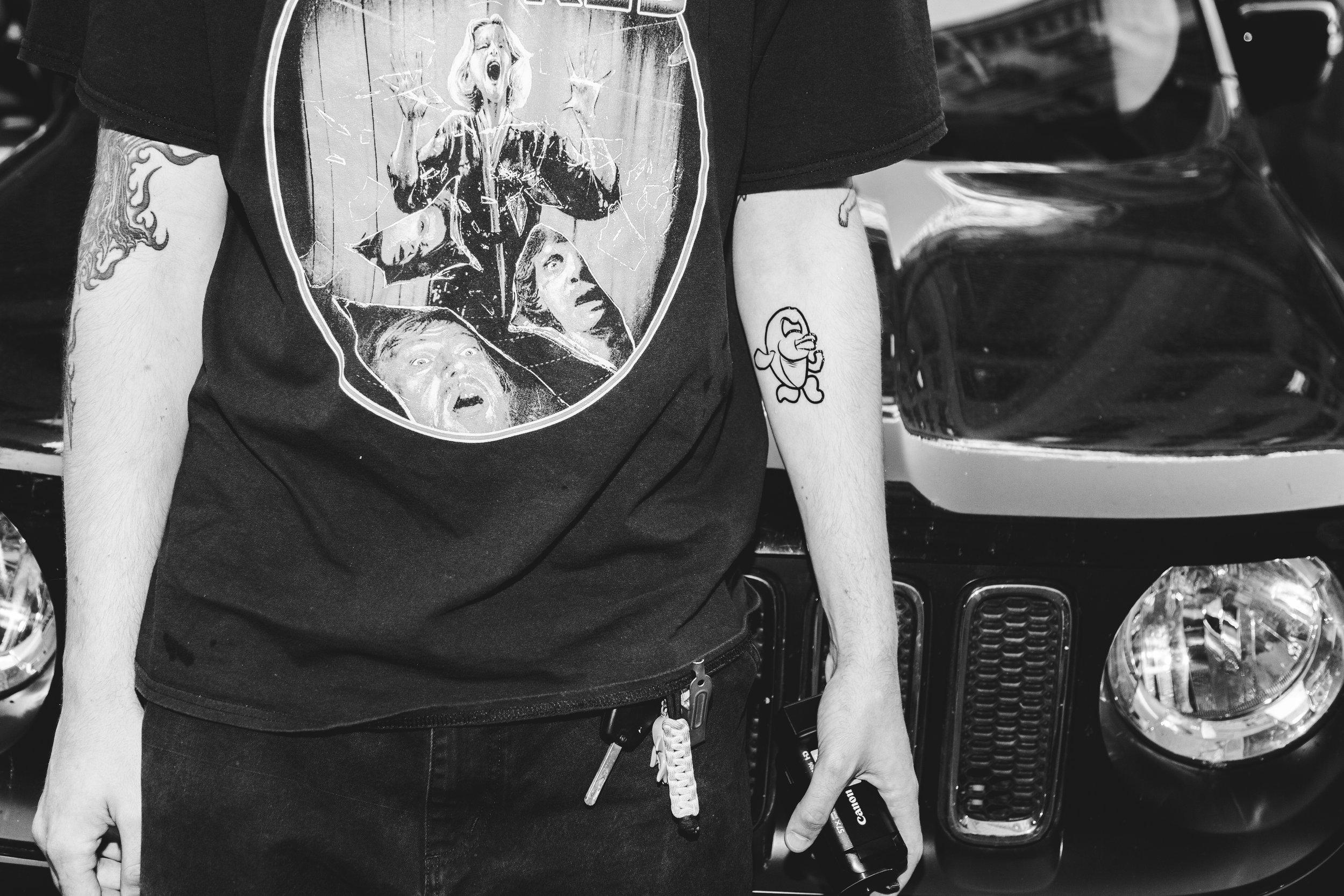 Person wearing a graphic T-shirt with horror movie characters, holding a backpack, with tattoos on arms, standing in front of a black vehicle.