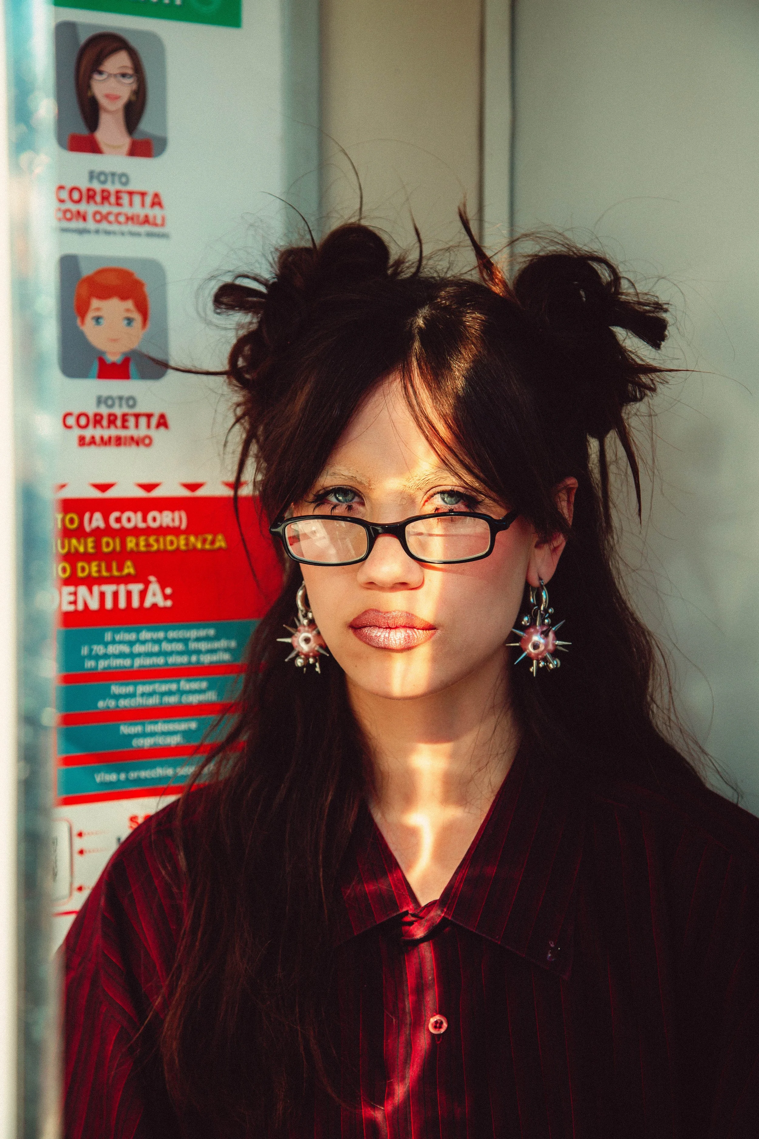 A woman with long, dark hair styled in a messy, spiky fashion, wearing glasses, earrings with starlike spikes, and a red, pinstripe shirt. She is standing in front of a sign with cartoon images and Italian text, and sunlight is casting shadows on her