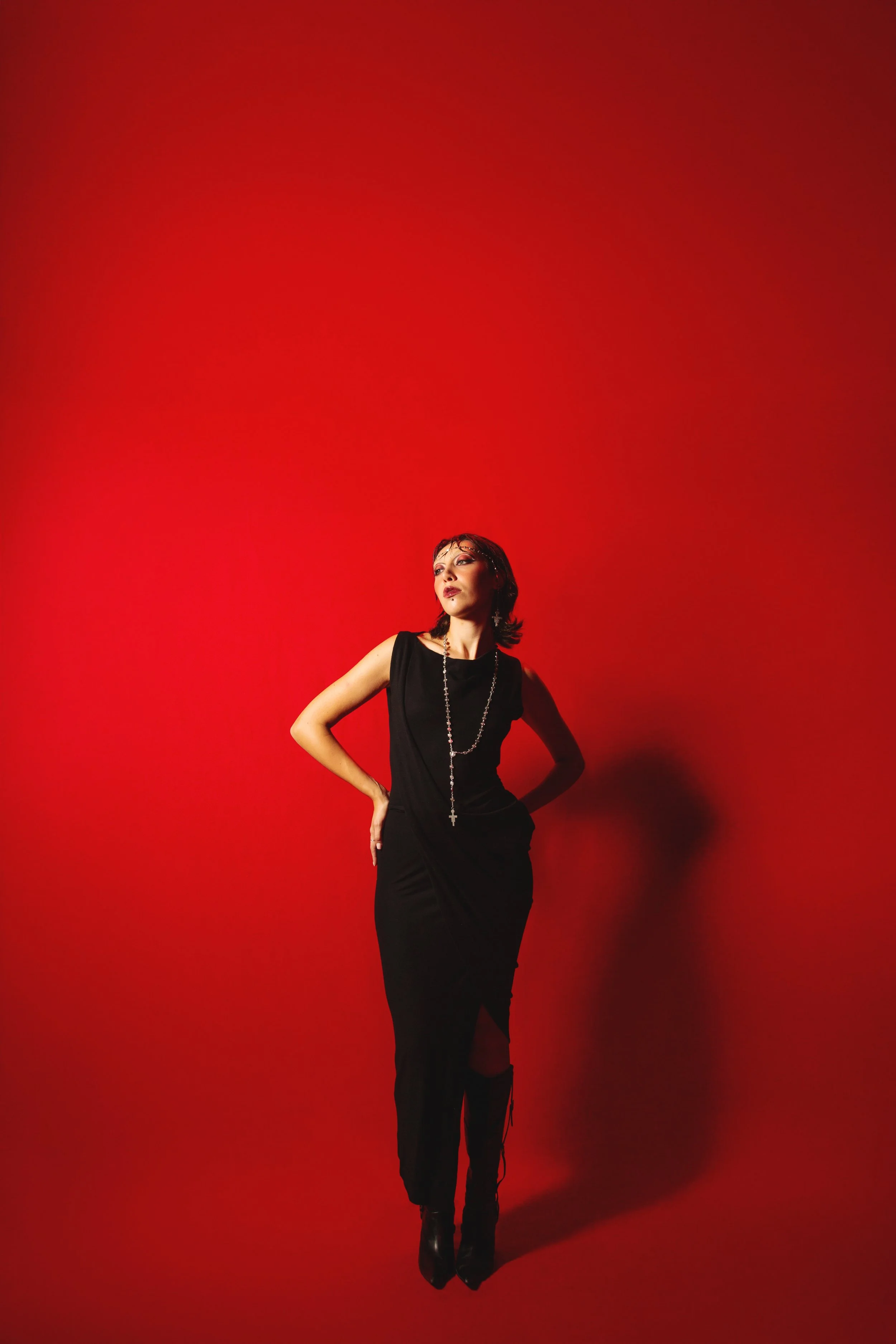 A woman dressed in black standing against a red background, wearing a long black dress, black boots, with her hands on her hips, and accessories including a long necklace and earrings.