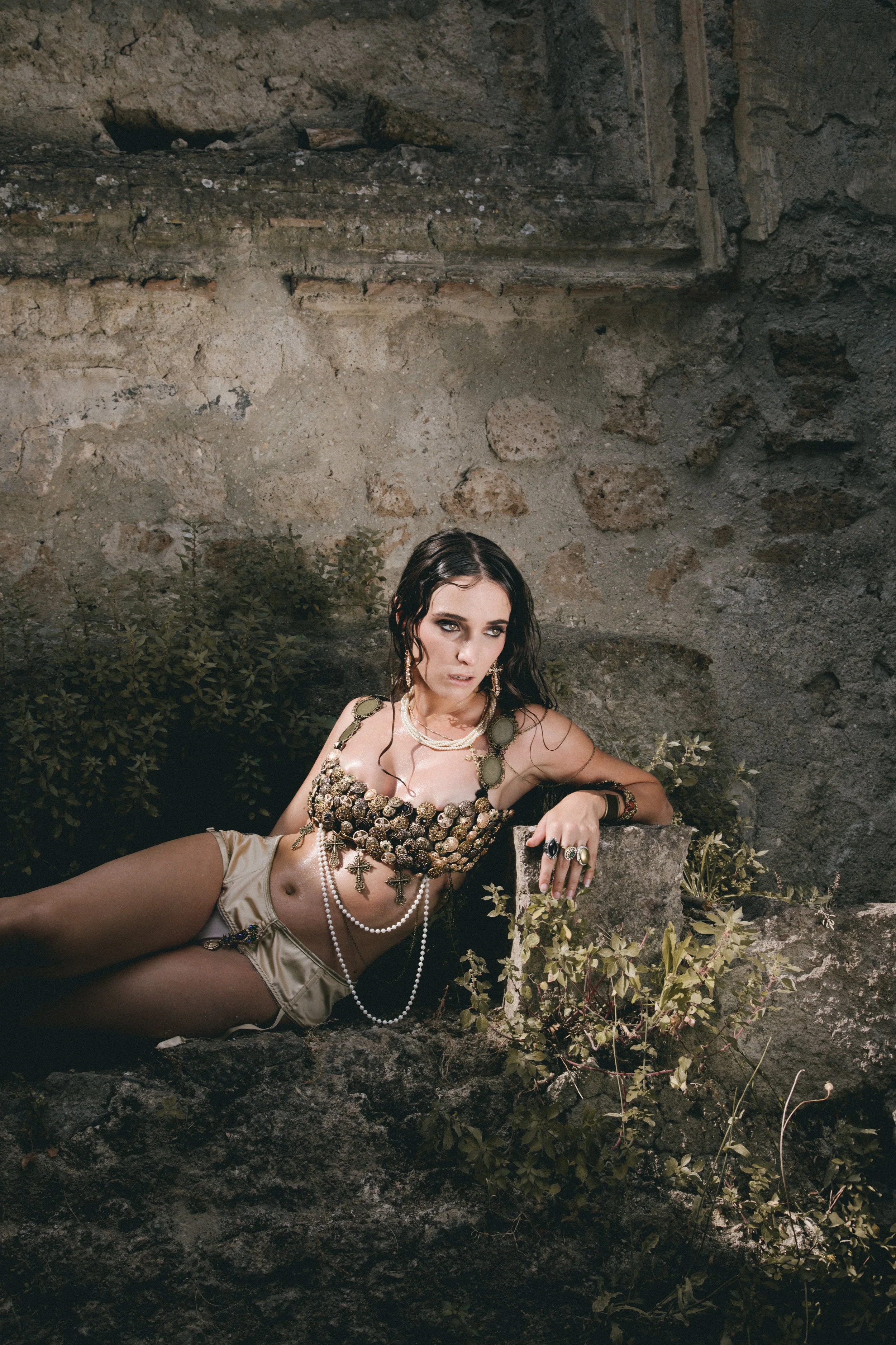 A woman with dark, wavy hair and jewelry, reclining against a stone ledge in front of a stone wall and greenery.