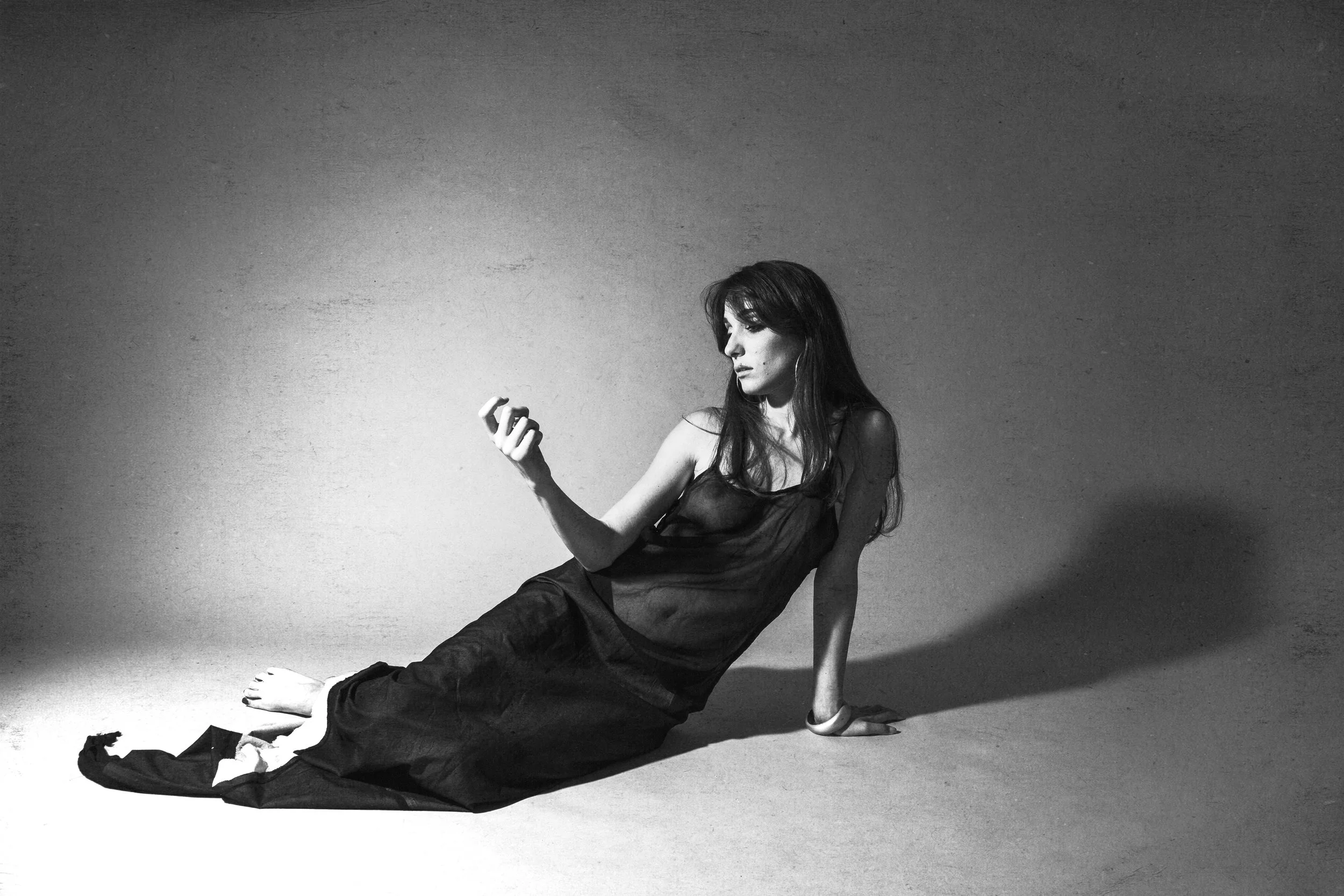 A woman sitting on the floor, dressed in a sheer black outfit, looking at her phone with a serious expression, in a studio setting with a plain background.