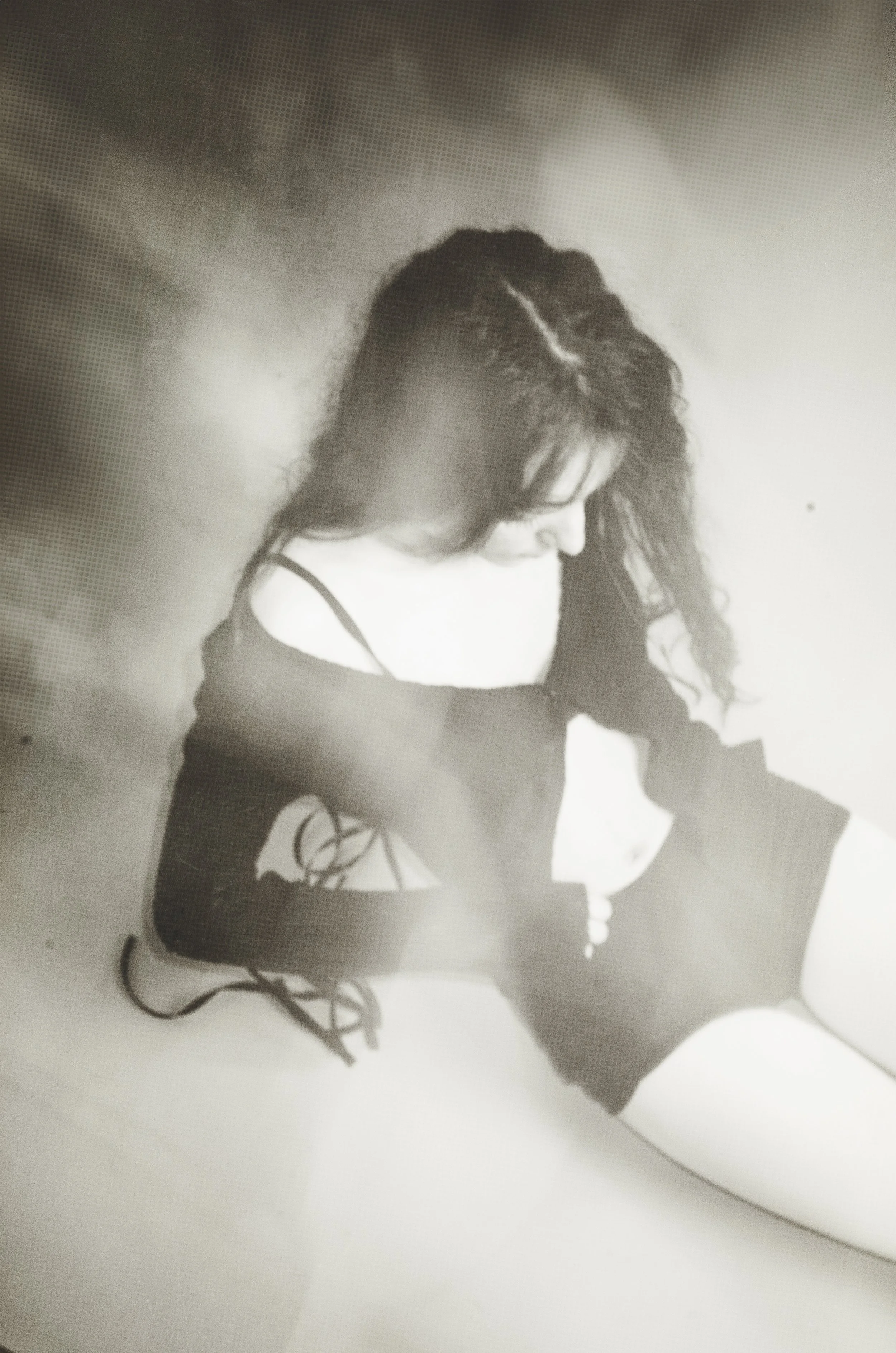 Black and white photo of a woman sitting on the floor with her head bowed, wearing a loose black top and shorts, with curly hair partially covering her face.