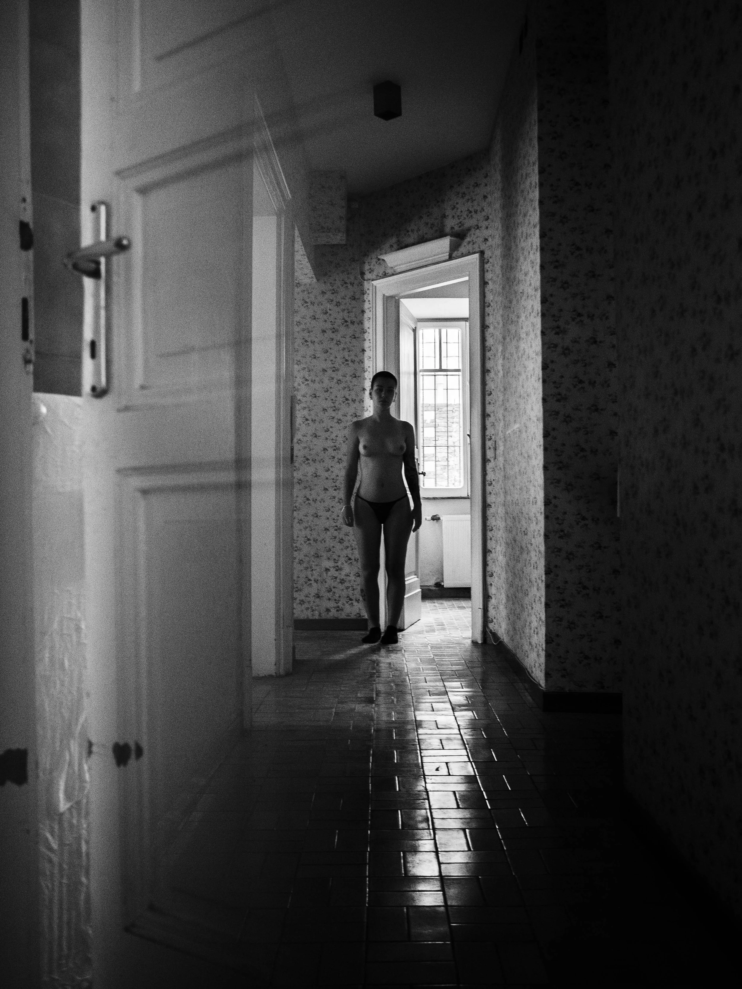 Black and white photo of a nude woman standing in a doorway, with the light coming from a window behind her, casting her silhouette.