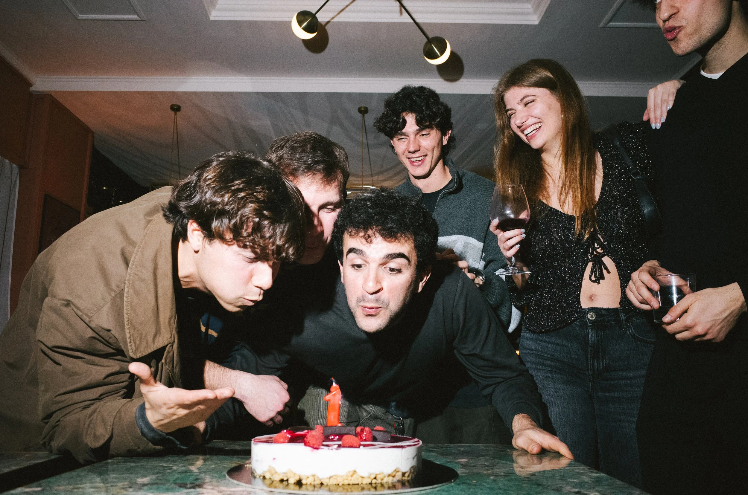 Group of friends celebrating a birthday, blowing out a single candle on a cake with raspberries, with some holding wine glasses, in a cozy indoor setting.