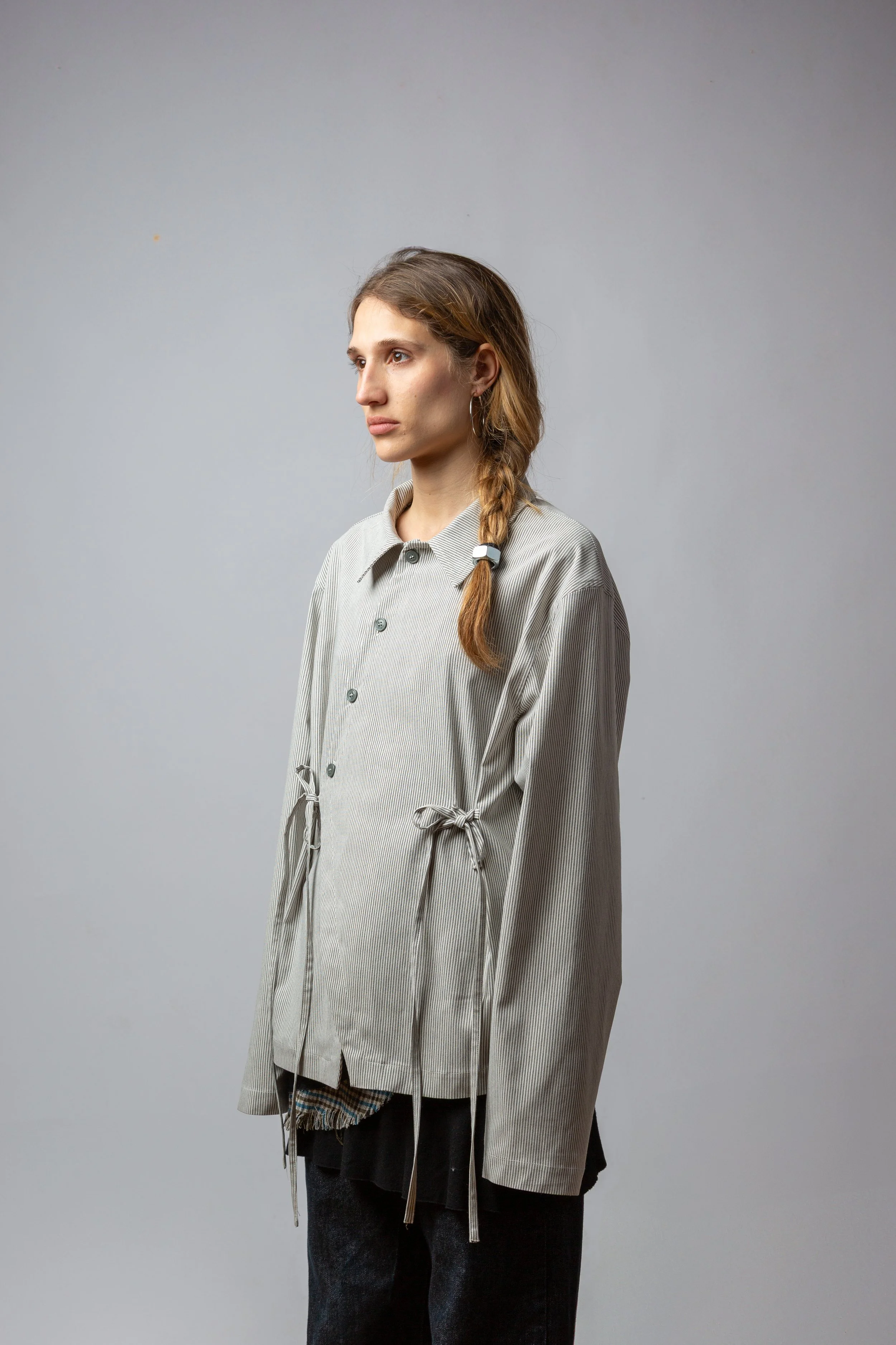 A young woman with long, wavy hair styled in a side braid, wearing a loose, grey striped button-up shirt with drawstrings on the sides and black pants, standing against a plain, light grey background.