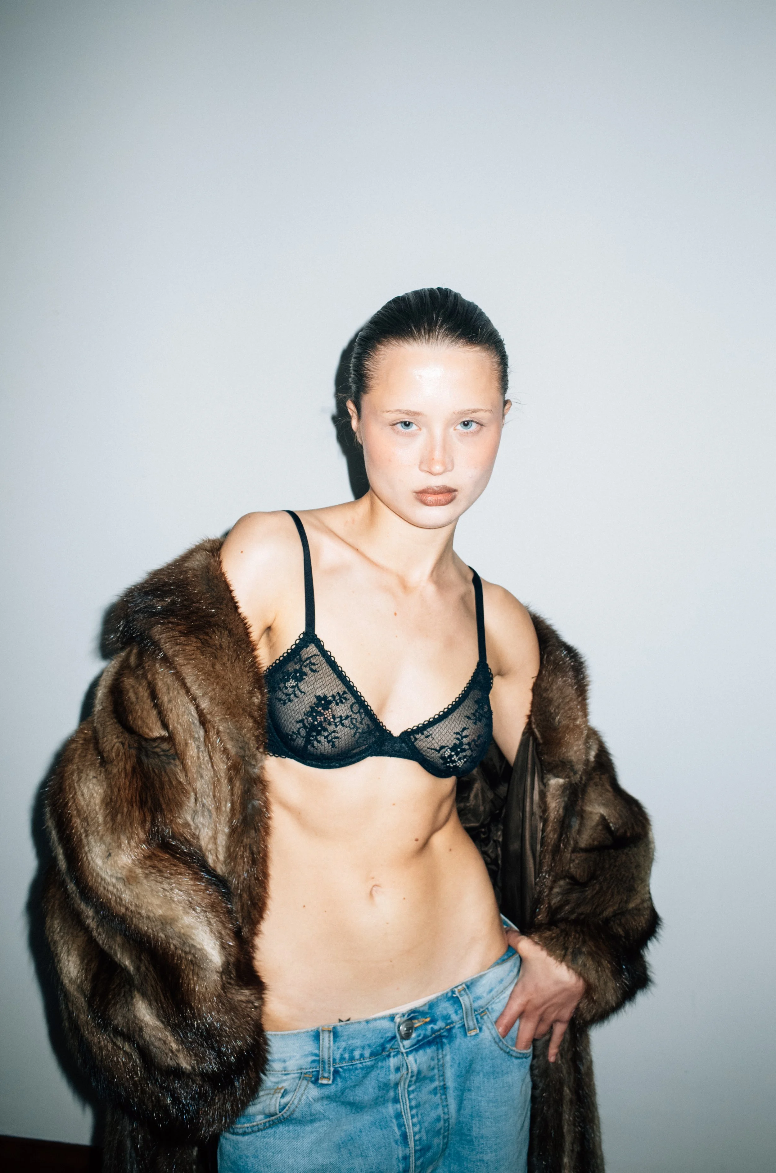 A woman with dark hair styled slicked back, wearing a sheer black lace bra, a fur coat draped over her shoulders, and blue jeans, standing against a plain light-colored wall.