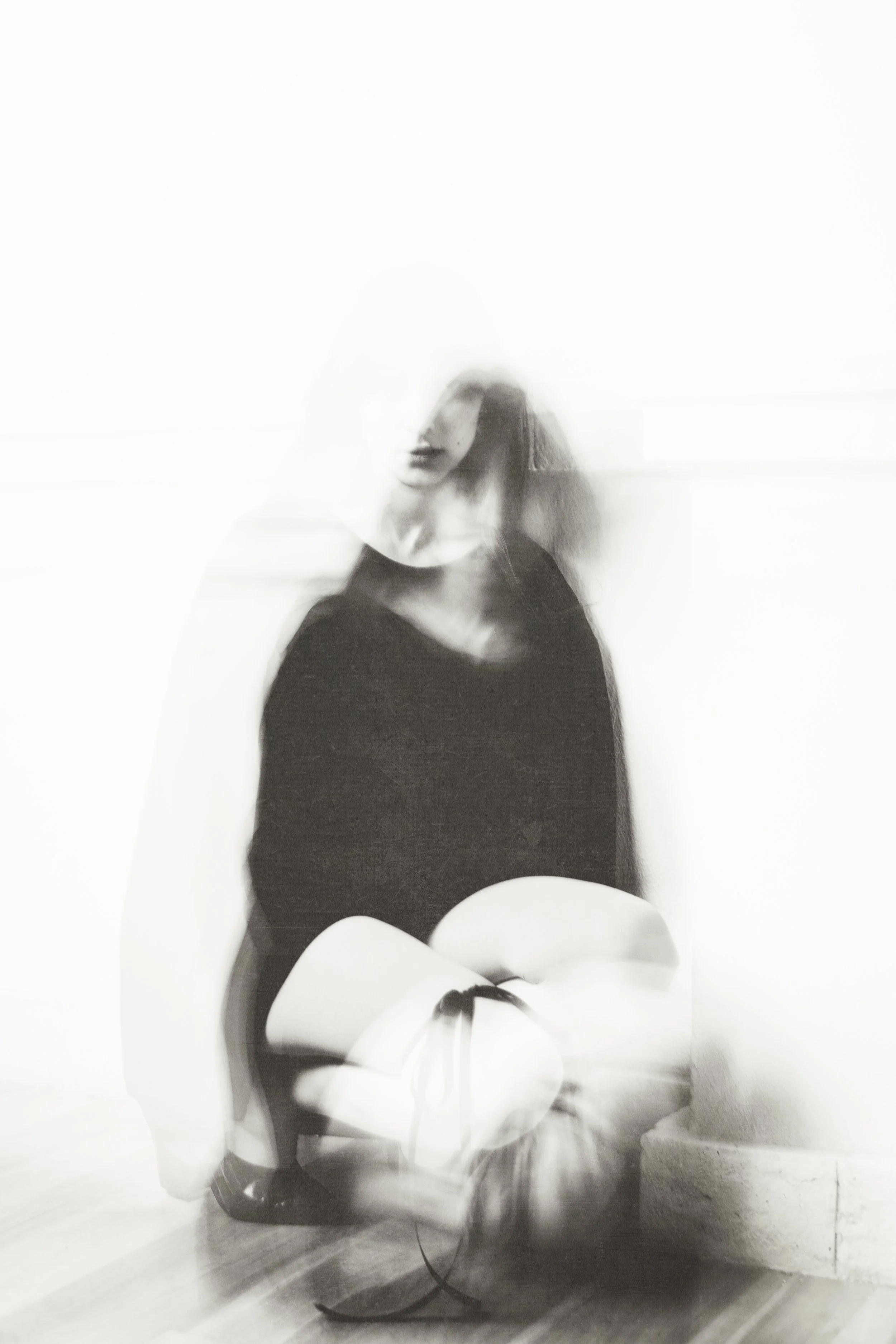 A black and white photograph of a woman with blurred movement, sitting on the floor with her legs crossed. She is wearing a dark top and has shoulder-length hair, with an abstract background.