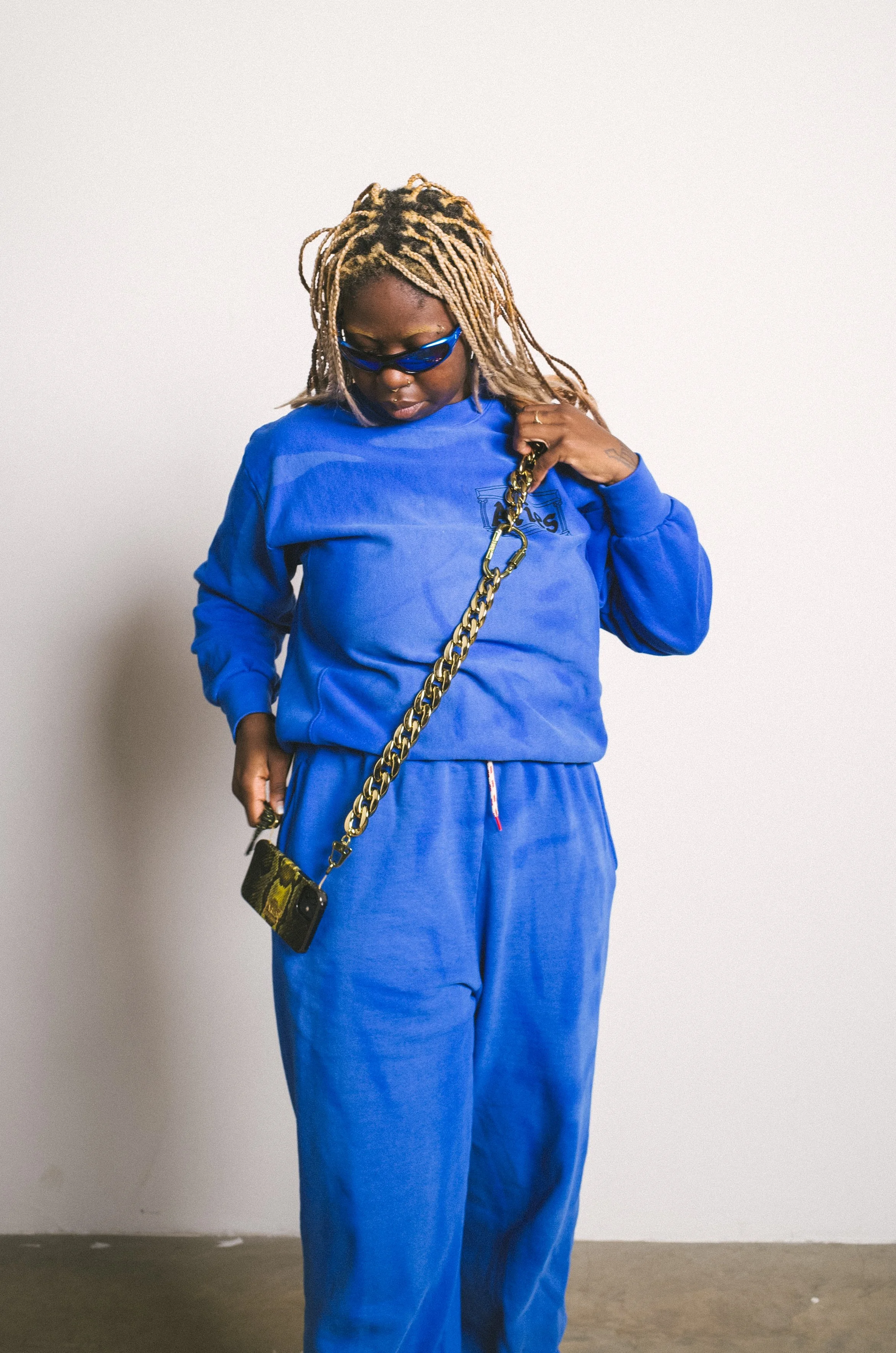 A woman with blonde dreadlocks wearing blue sunglasses, a blue sweatshirt, and matching blue pants, holding a small phone with a gold chain strap.