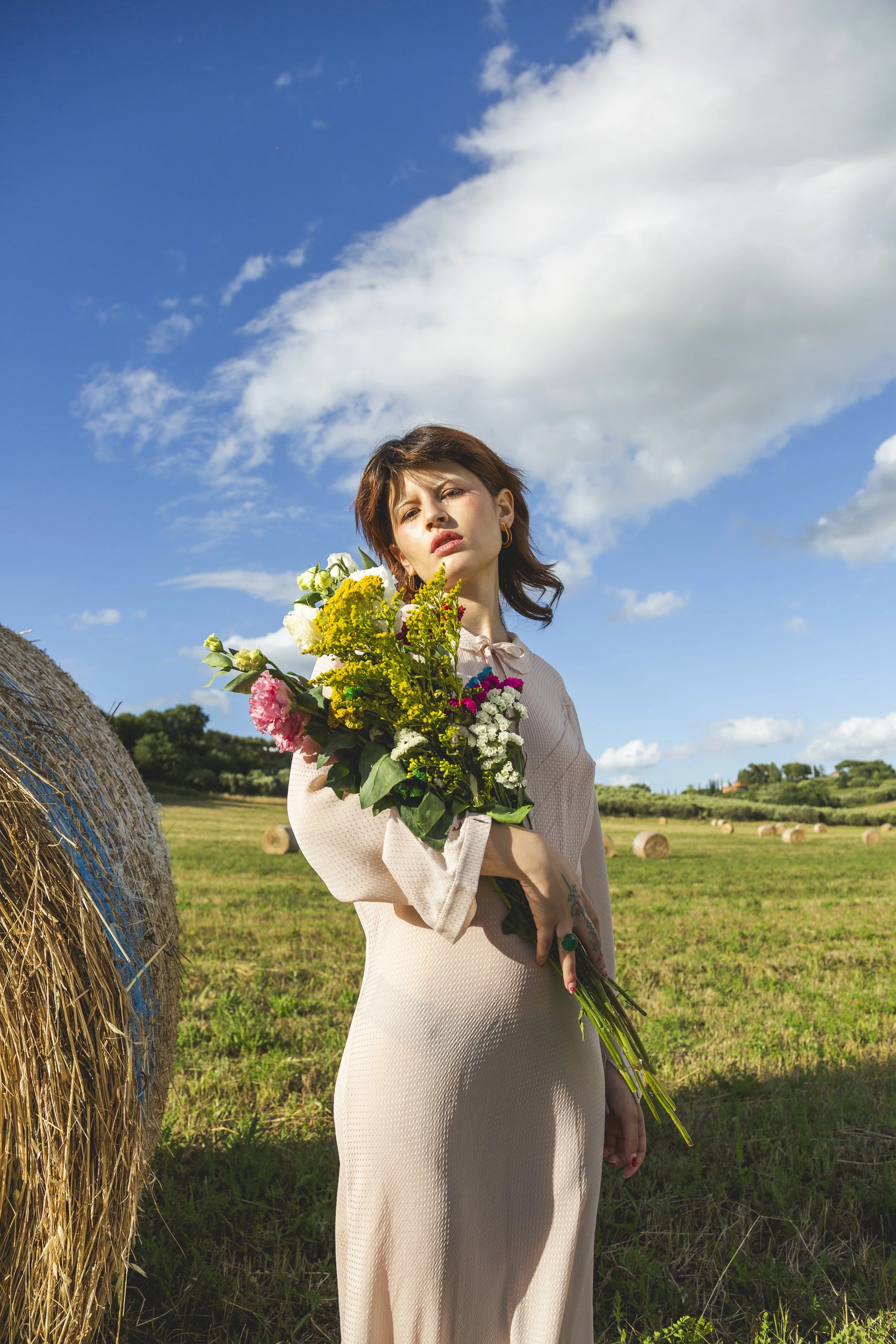 A woman standing in a field holding a bouquet of colorful flowers, with hay bales and a blue sky with clouds in the background.