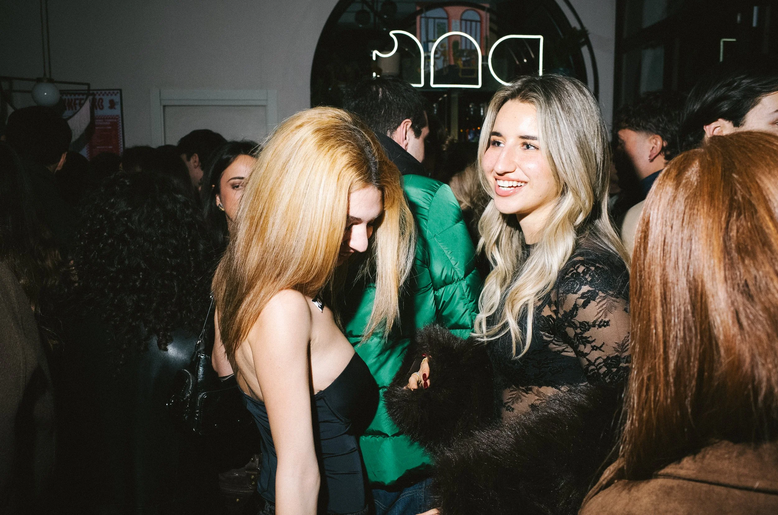 Two women are smiling and socializing at a crowded party or club. One woman has long blonde hair and is wearing a black lace dress, while the other woman has light blonde hair and is wearing a black dress with fuzzy black gloves.