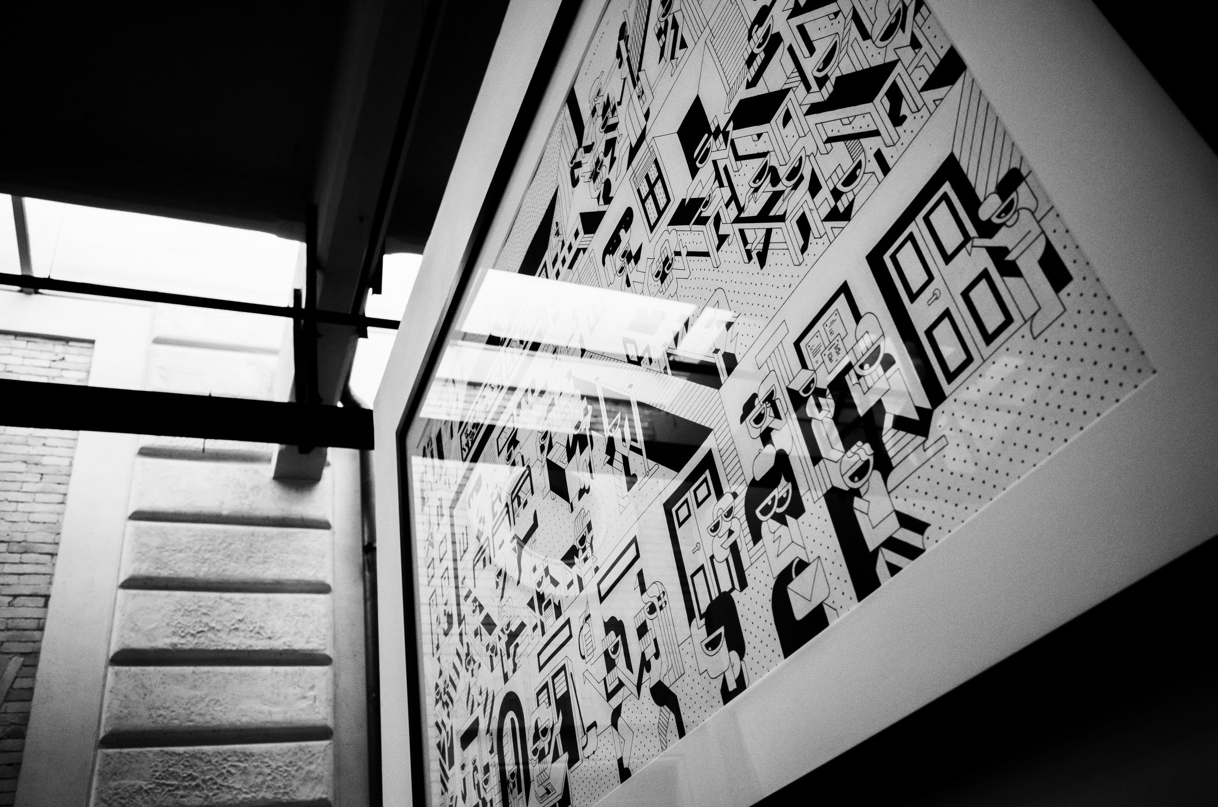 An abstract mural with black and white geometric patterns and illustrations, seen from below at an angle, next to a brick wall and large window.
