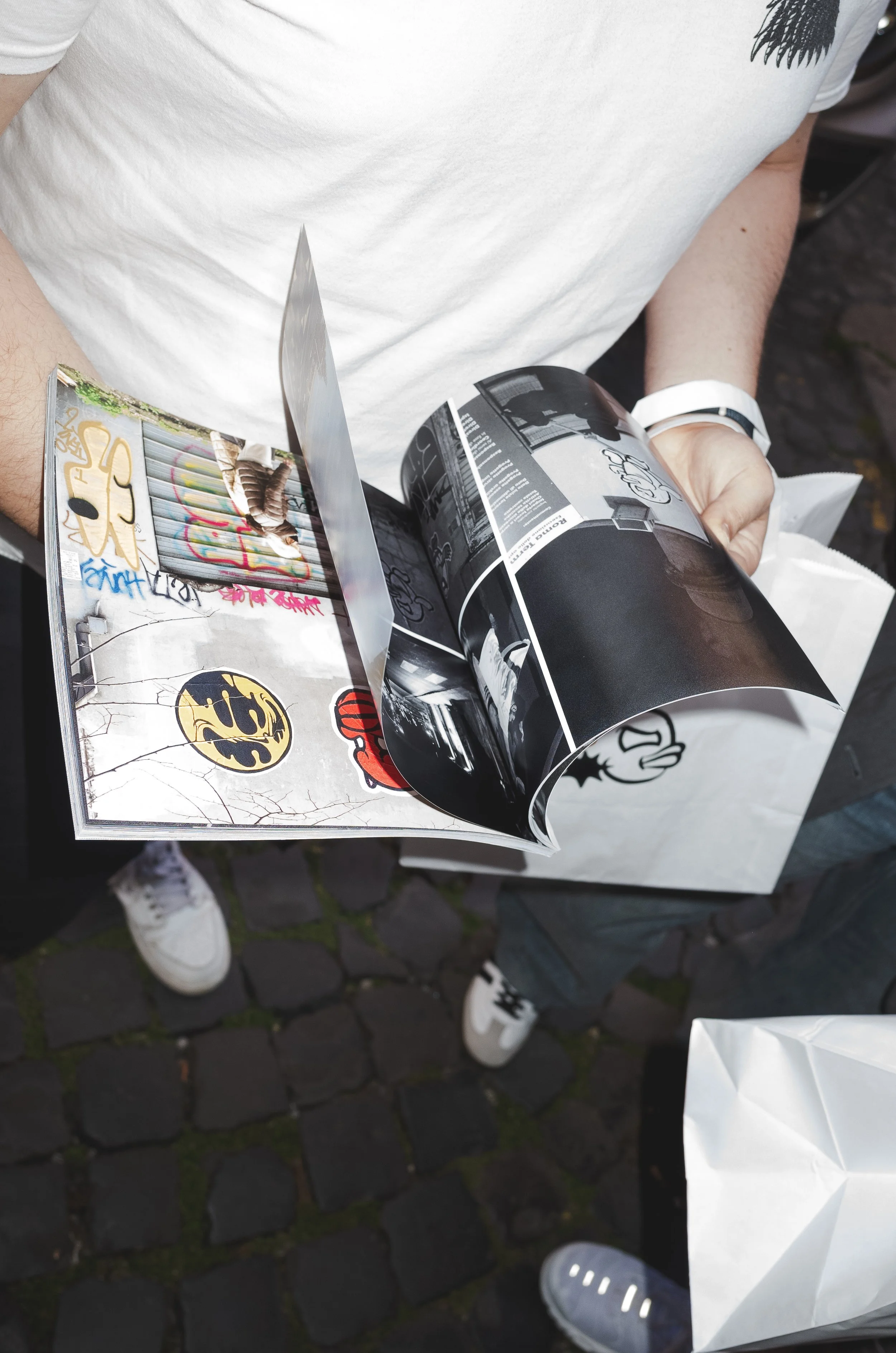 Person holding a magazine with photos and graffiti on the cover, standing on cobblestone street.