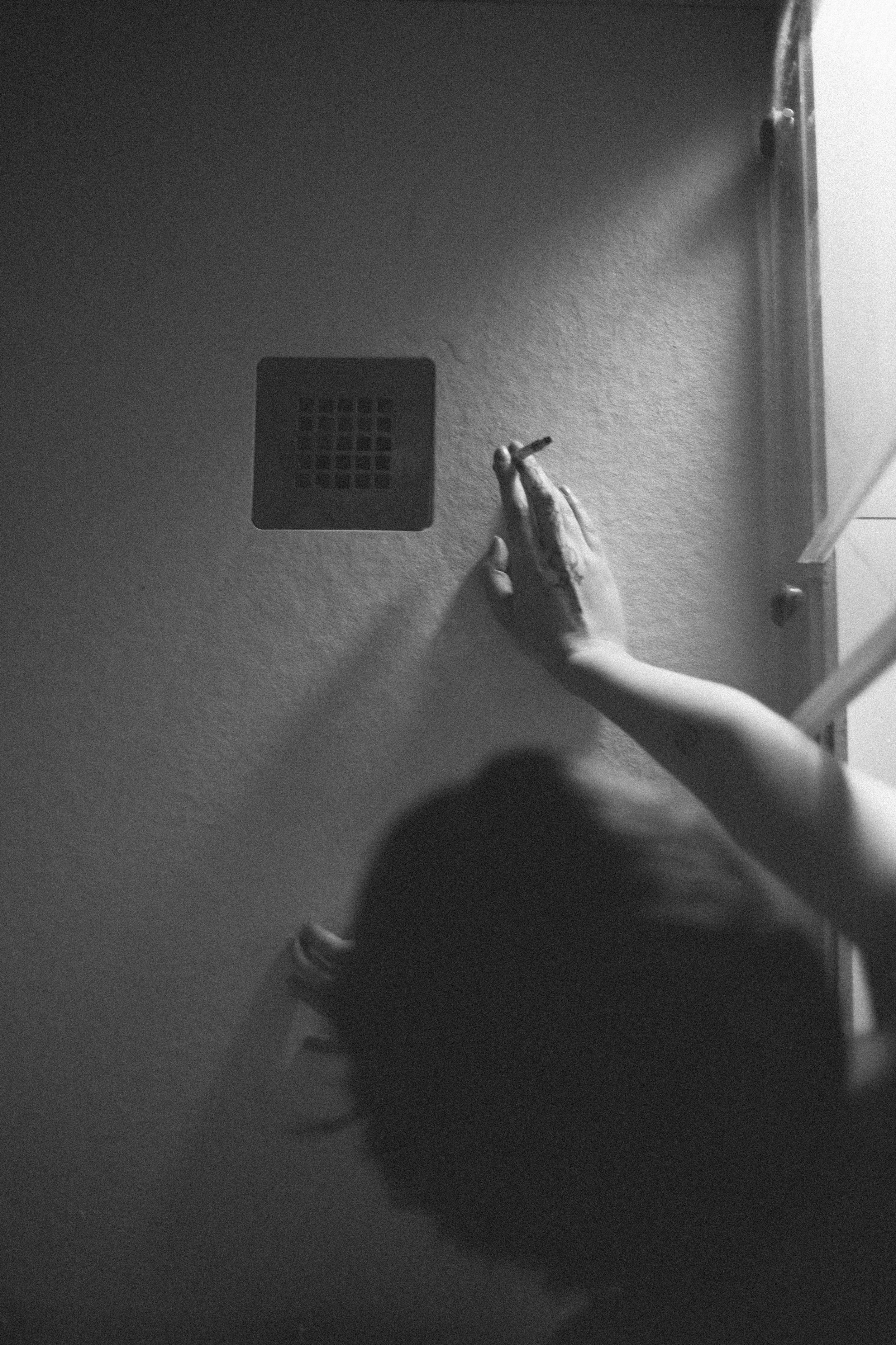 A black and white photo from above showing a person with long hair and a tattoo on their hand, reaching towards a ceiling vent on a wall, with part of their head and arm visible.