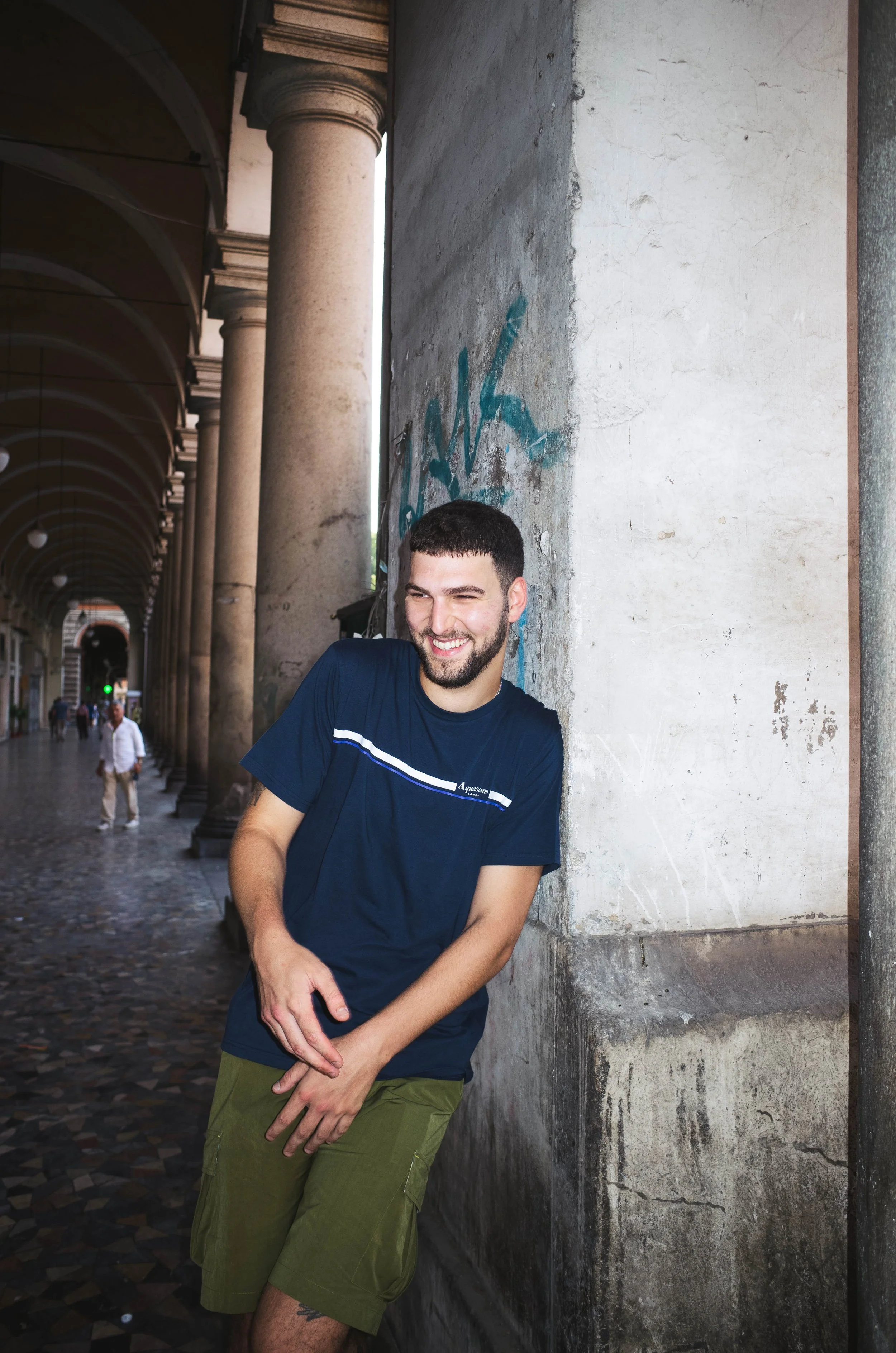 A smiling man with a beard and short dark hair, wearing a dark blue t-shirt and olive green shorts, leaning against a concrete wall with graffiti in an outdoor corridor with columns.