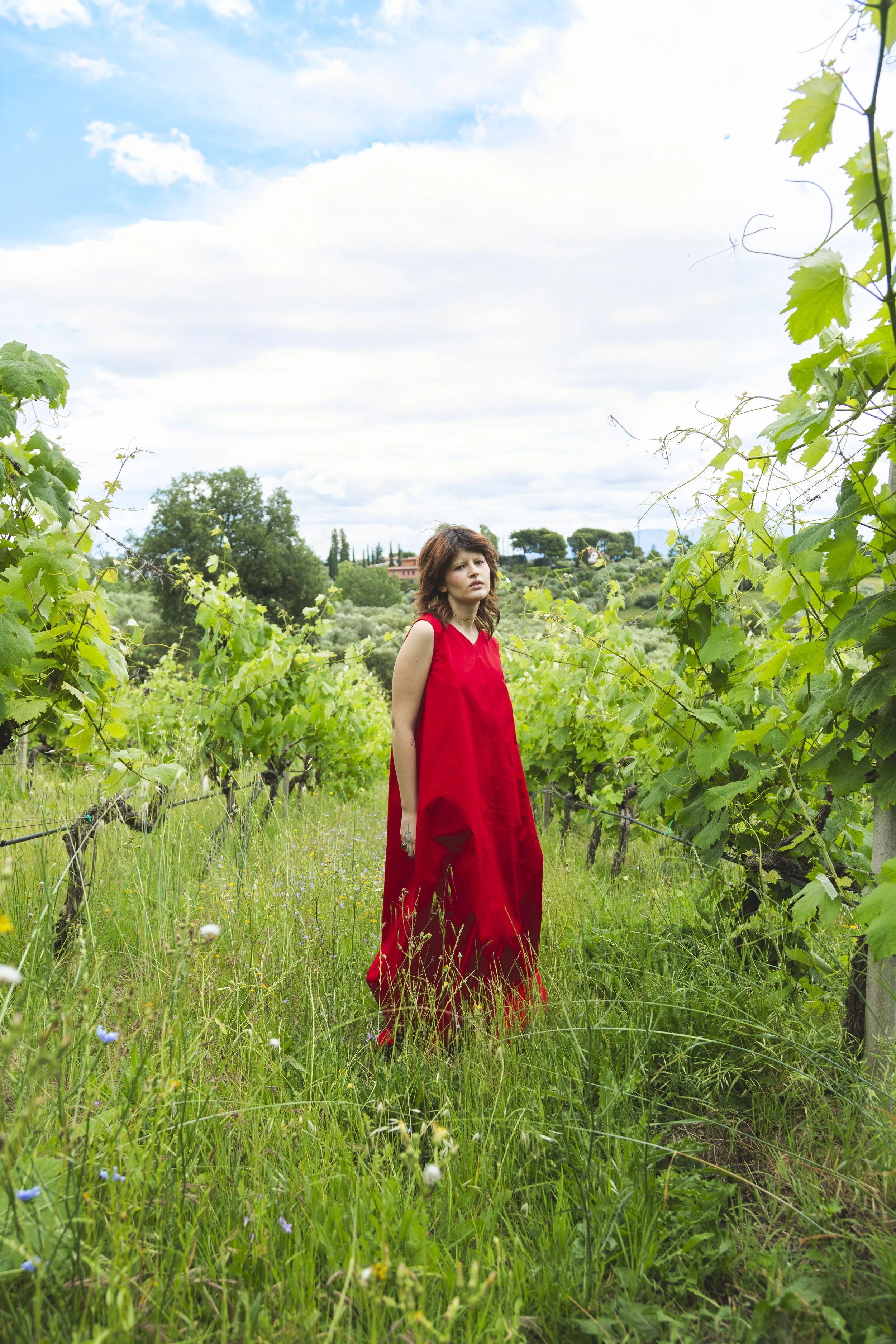 A woman in a long red dress stands in a vineyard with green grapevines and a lush landscape under a partly cloudy sky.