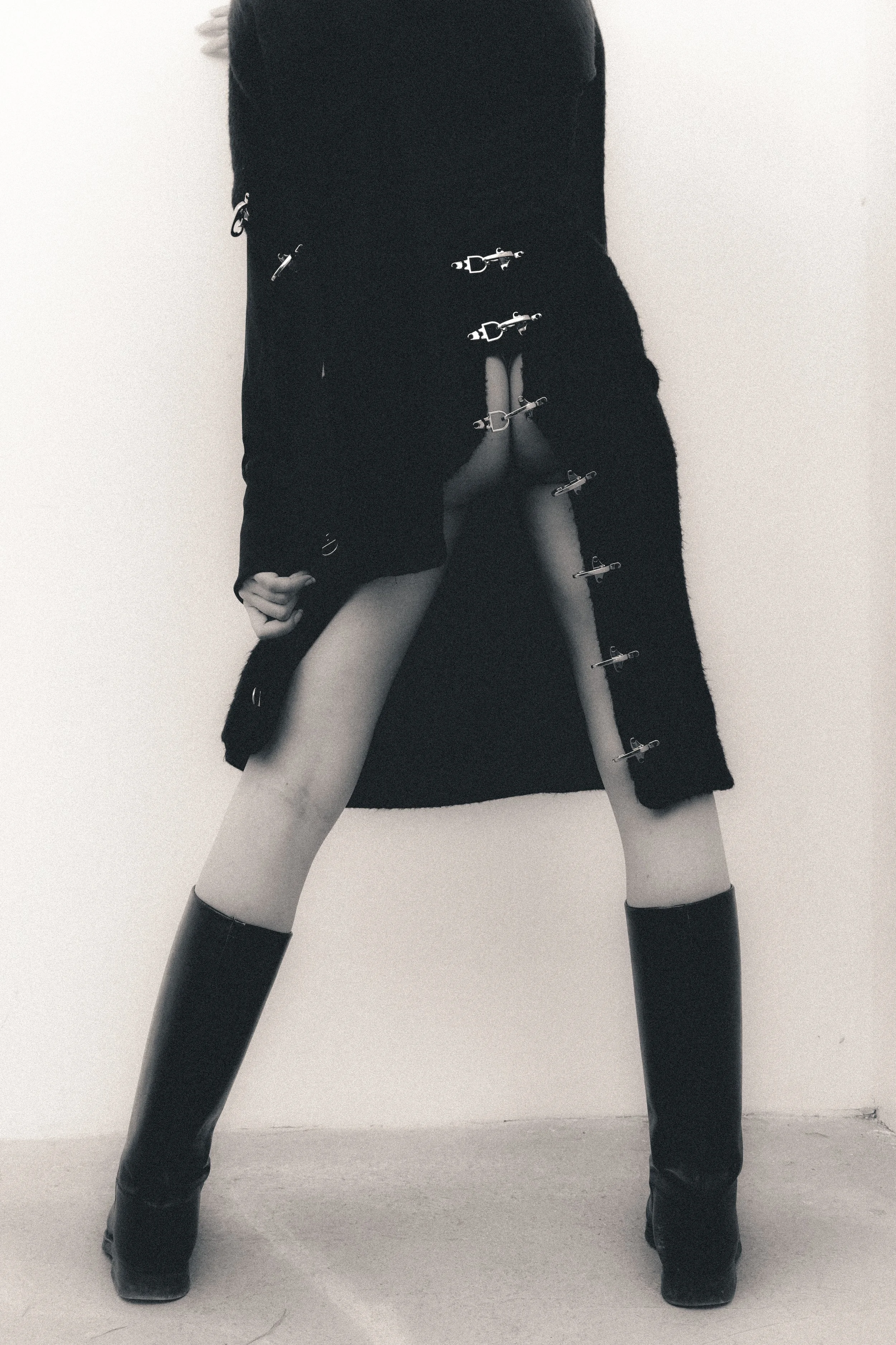 A person in black boots and a long black coat with metal clips, standing against a plain wall, with legs apart and slightly bent, in a provocative pose.