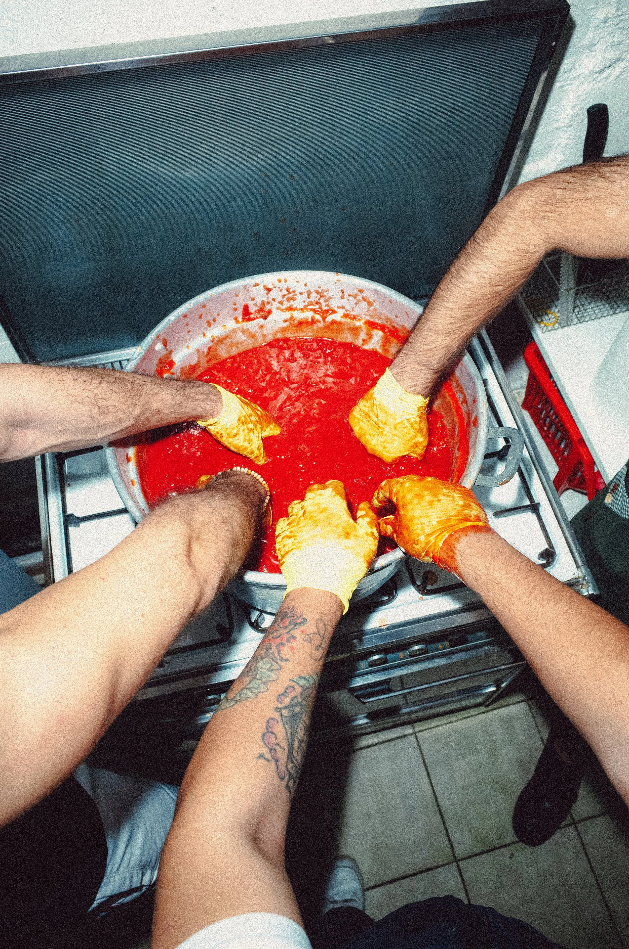 Three people stirring a large pot of red marinara sauce on a kitchen stove, wearing yellow gloves.
