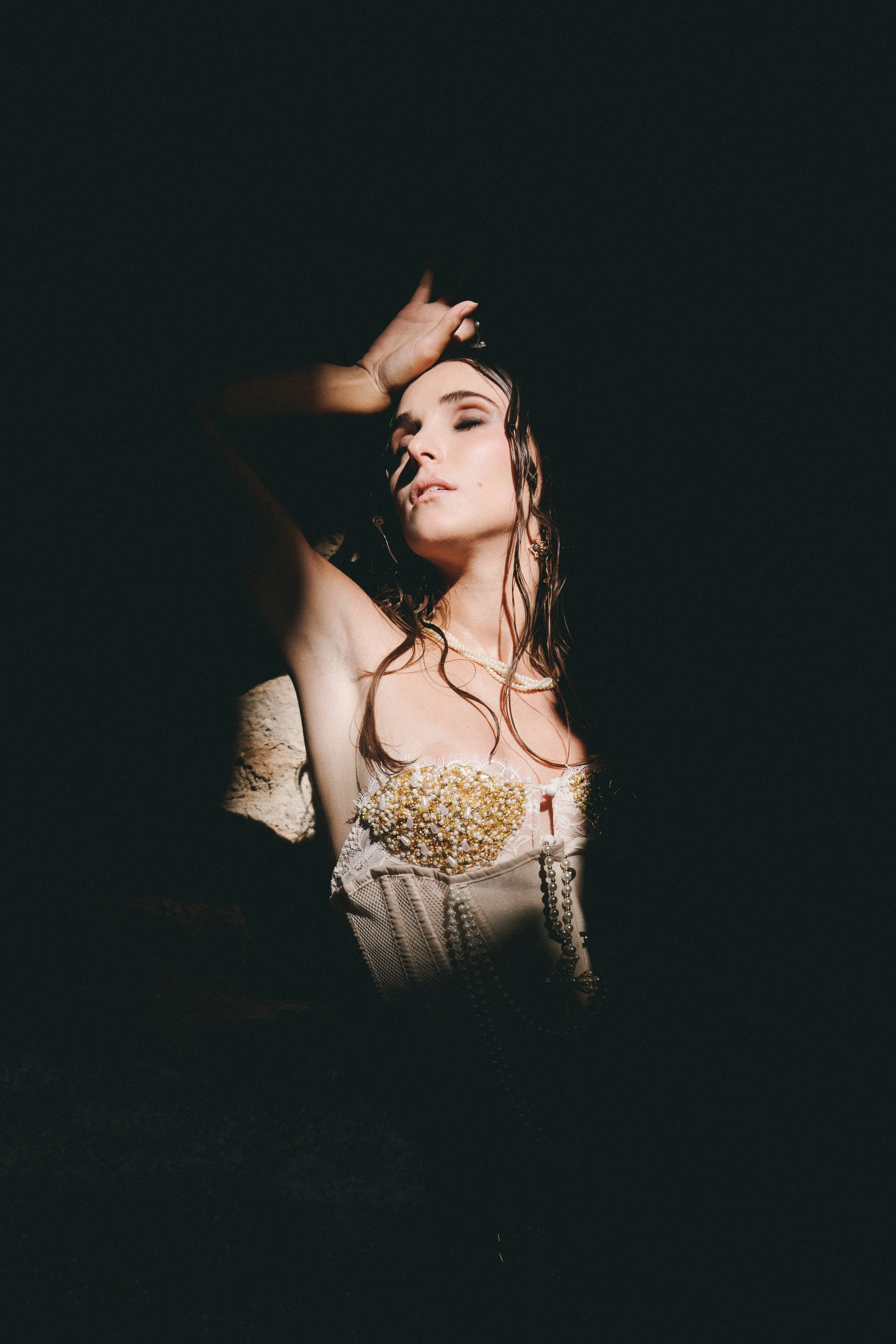 A woman with wet hair and jewelry, posing with her eyes closed and hand on her forehead, against a dark background.