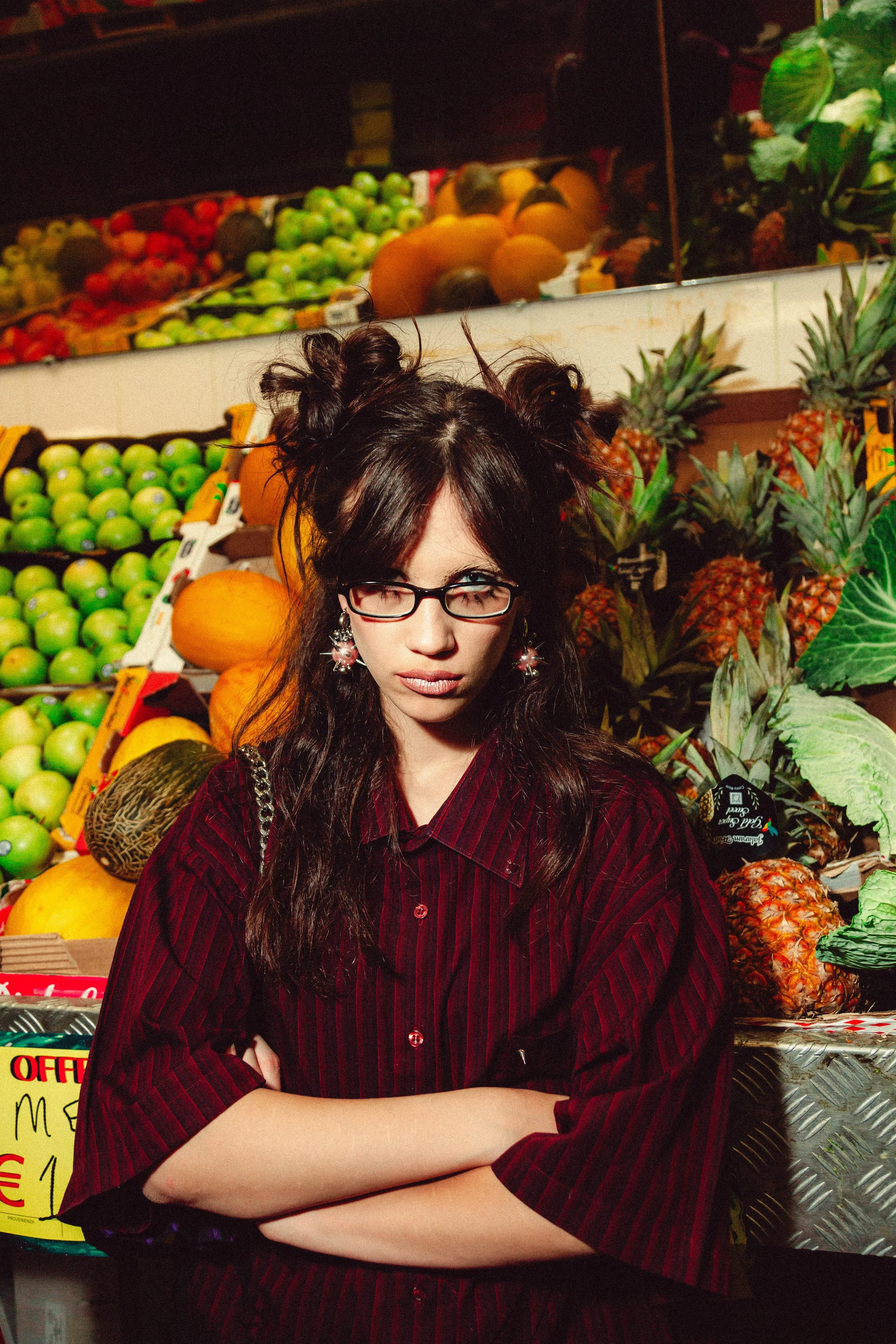 A woman with glasses and earrings standing in front of a produce stand with fruits including apples, pineapples, and pumpkins.