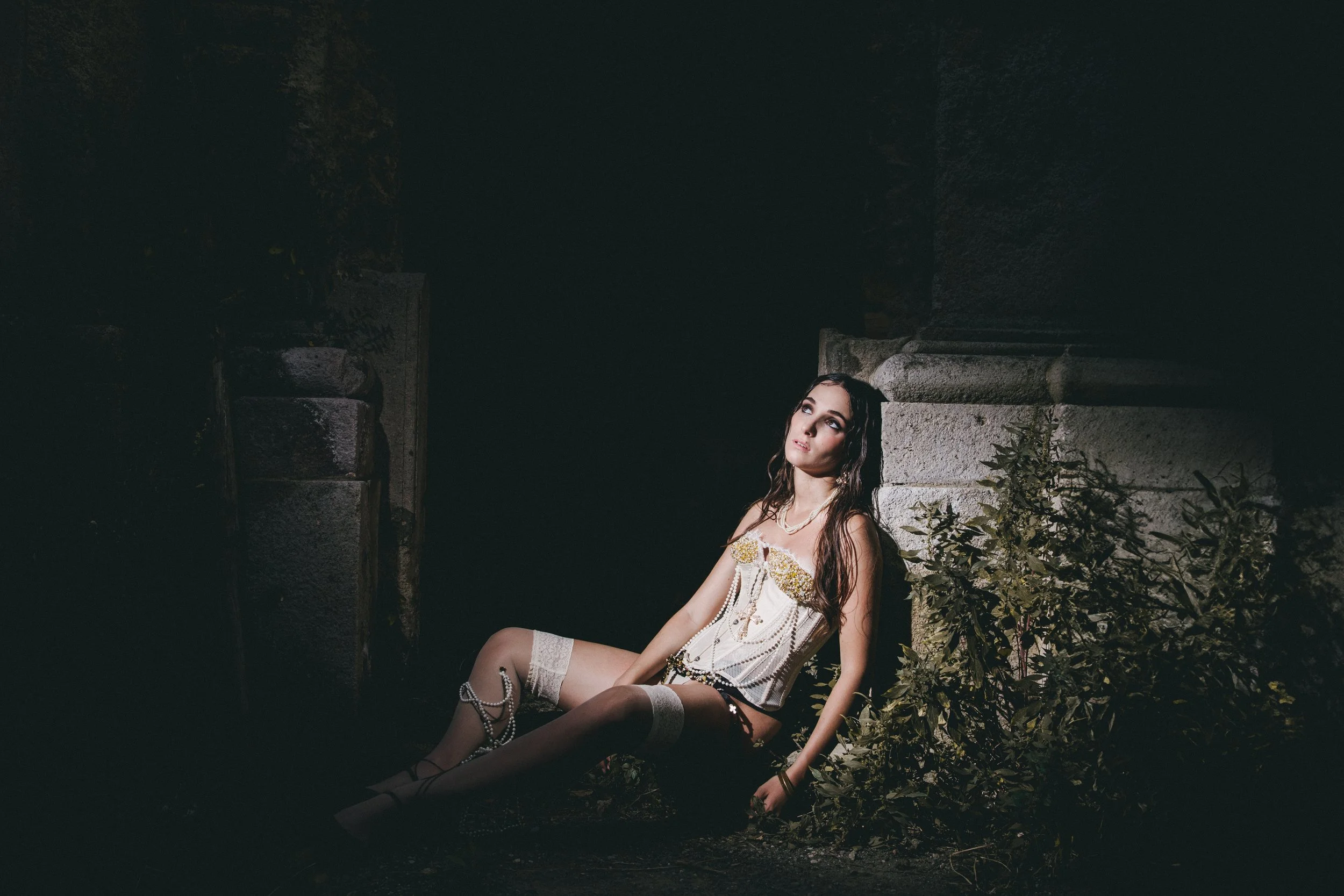 A woman in vintage lingerie sitting on the ground near stone ruins at night, illuminated by a focused light.