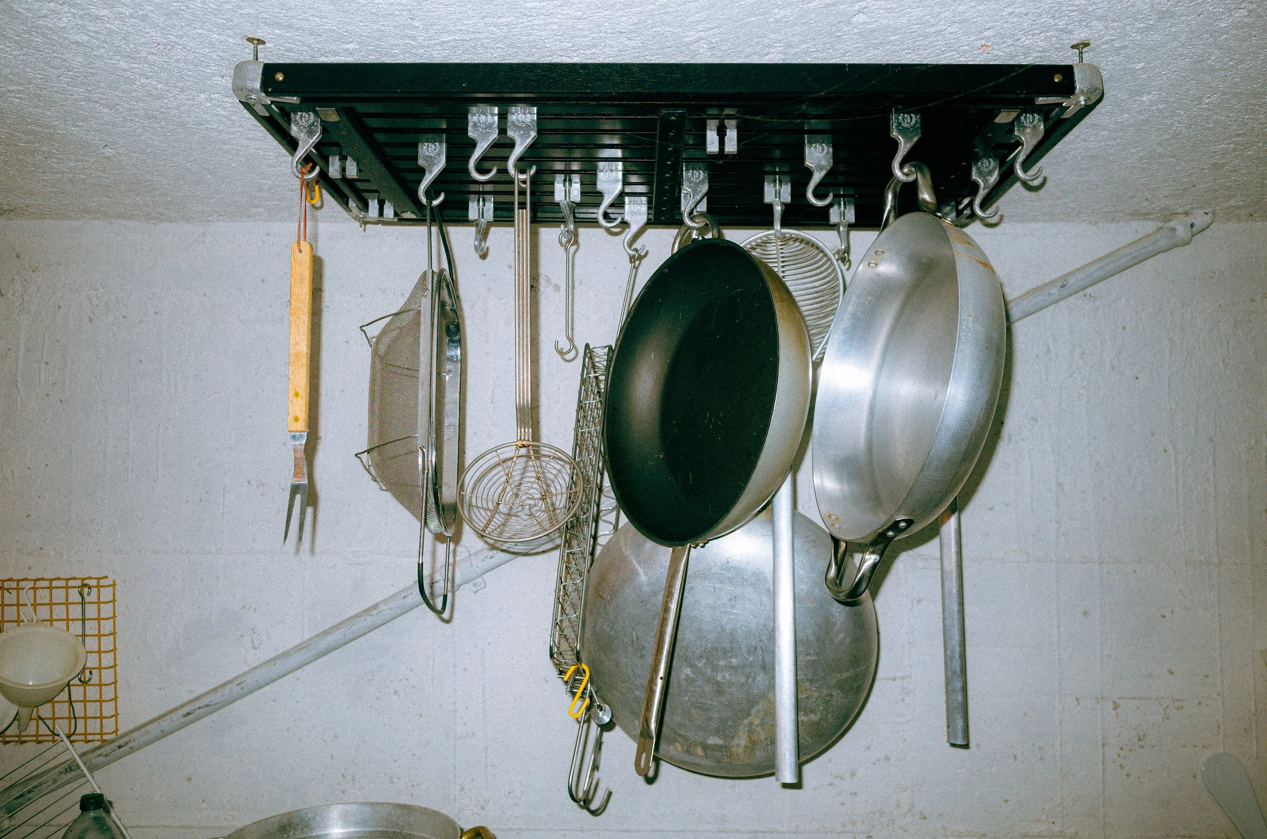 Hanging kitchen utensils and cookware, including pots, pans, colanders, and a cooking fork, on a ceiling rack.