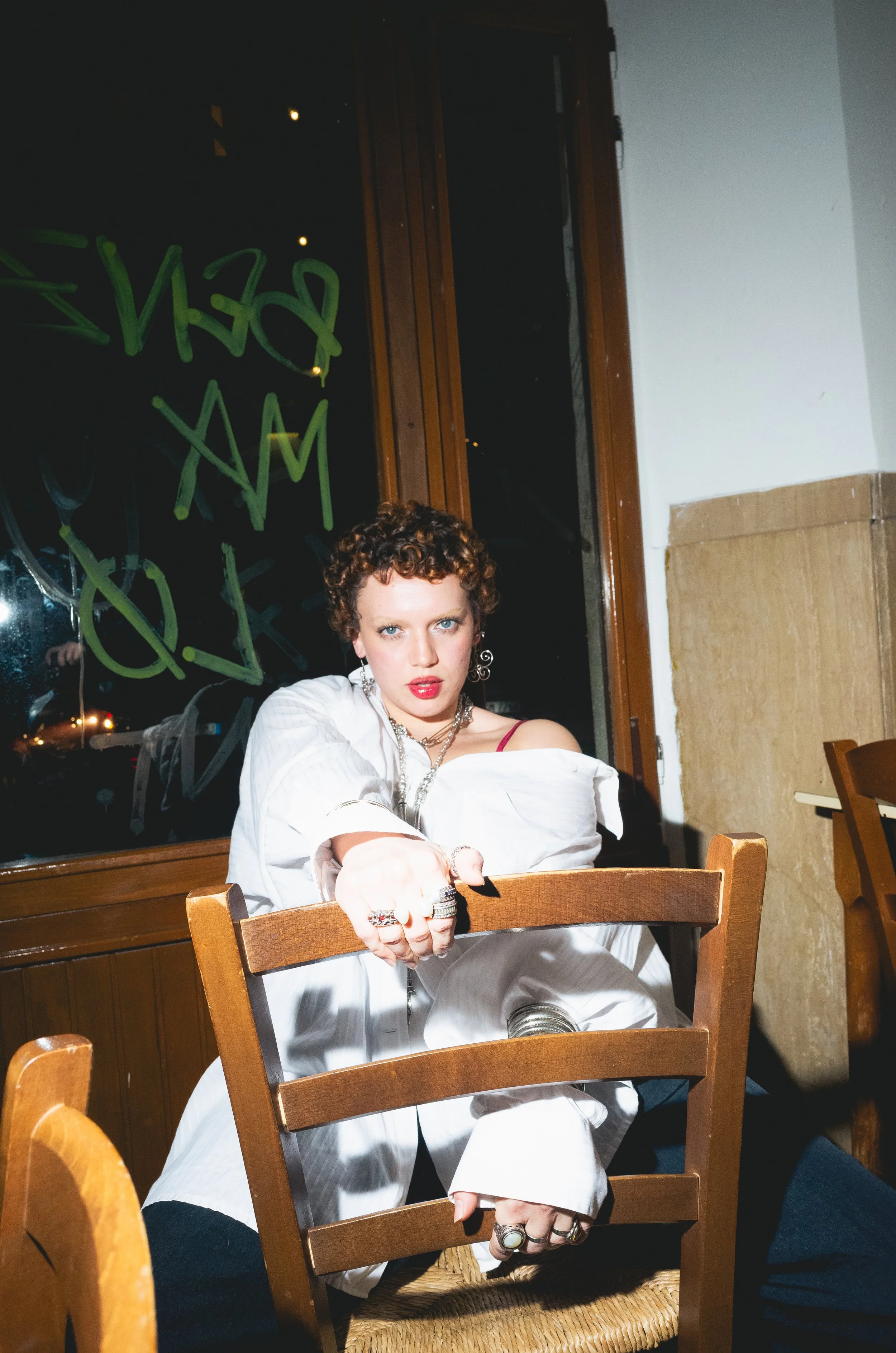 A woman with short, curly hair and bold makeup is sitting at a table in a restaurant, leaning forward with her arm resting on a wooden chair, looking directly at the camera with a confident expression, wearing a white shirt, jewelry, and a red strap 