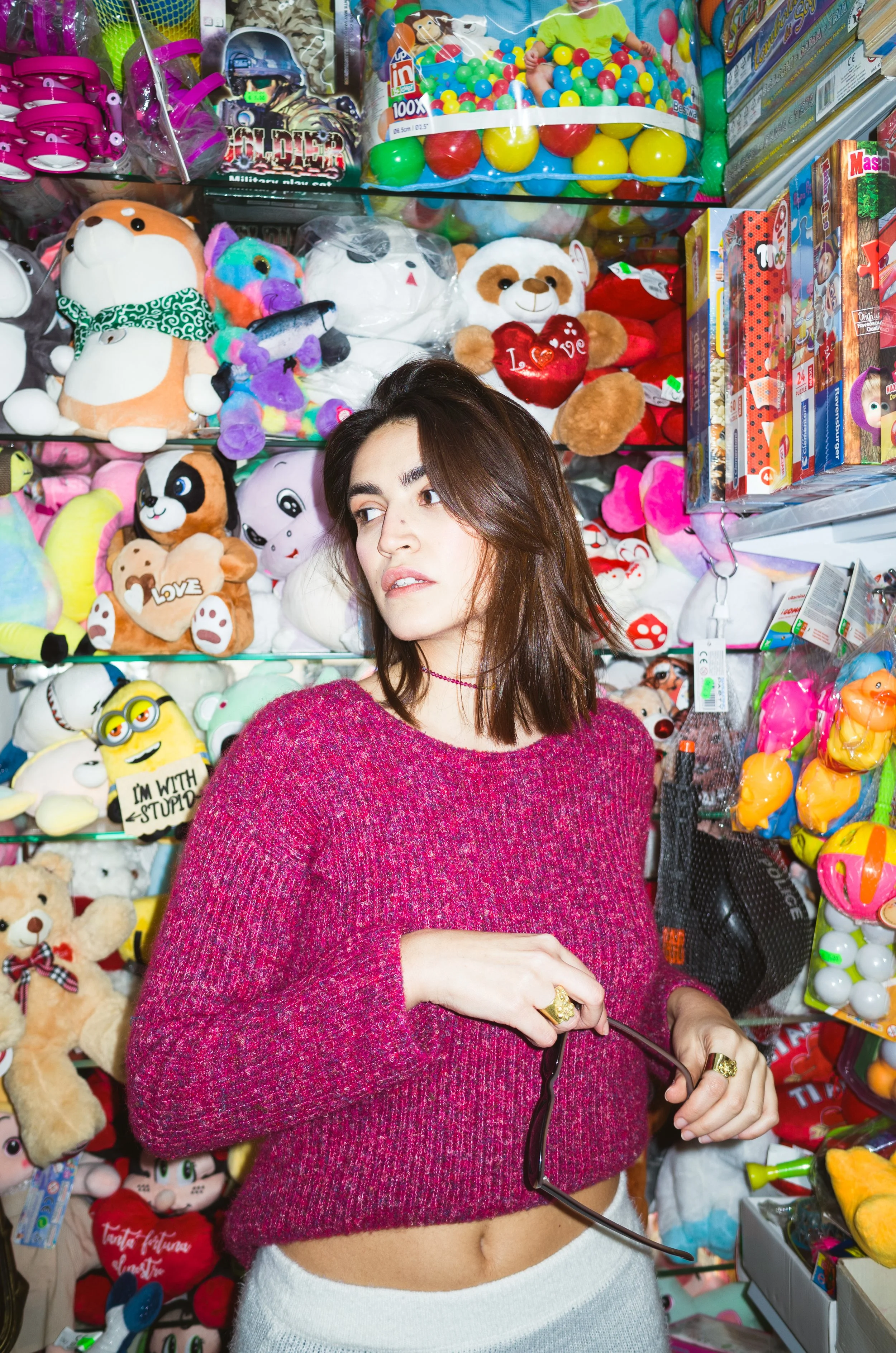 A woman in a pink sweater and light-colored pants stands in front of a wall of stuffed animals and toys in a store, holding sunglasses.