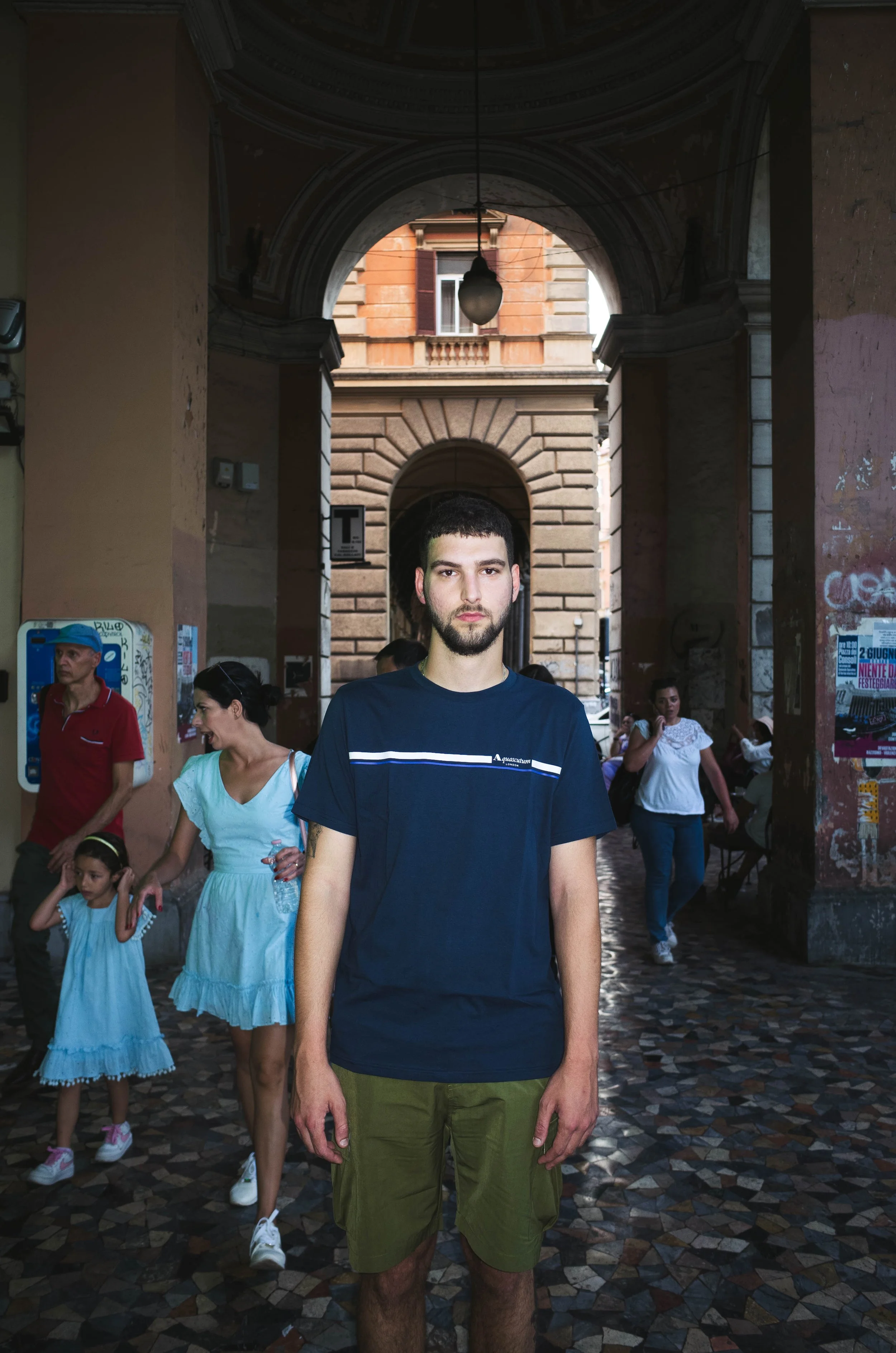 Young man with dark hair and beard standing in the center of a busy street under an archway, with people and buildings in the background.