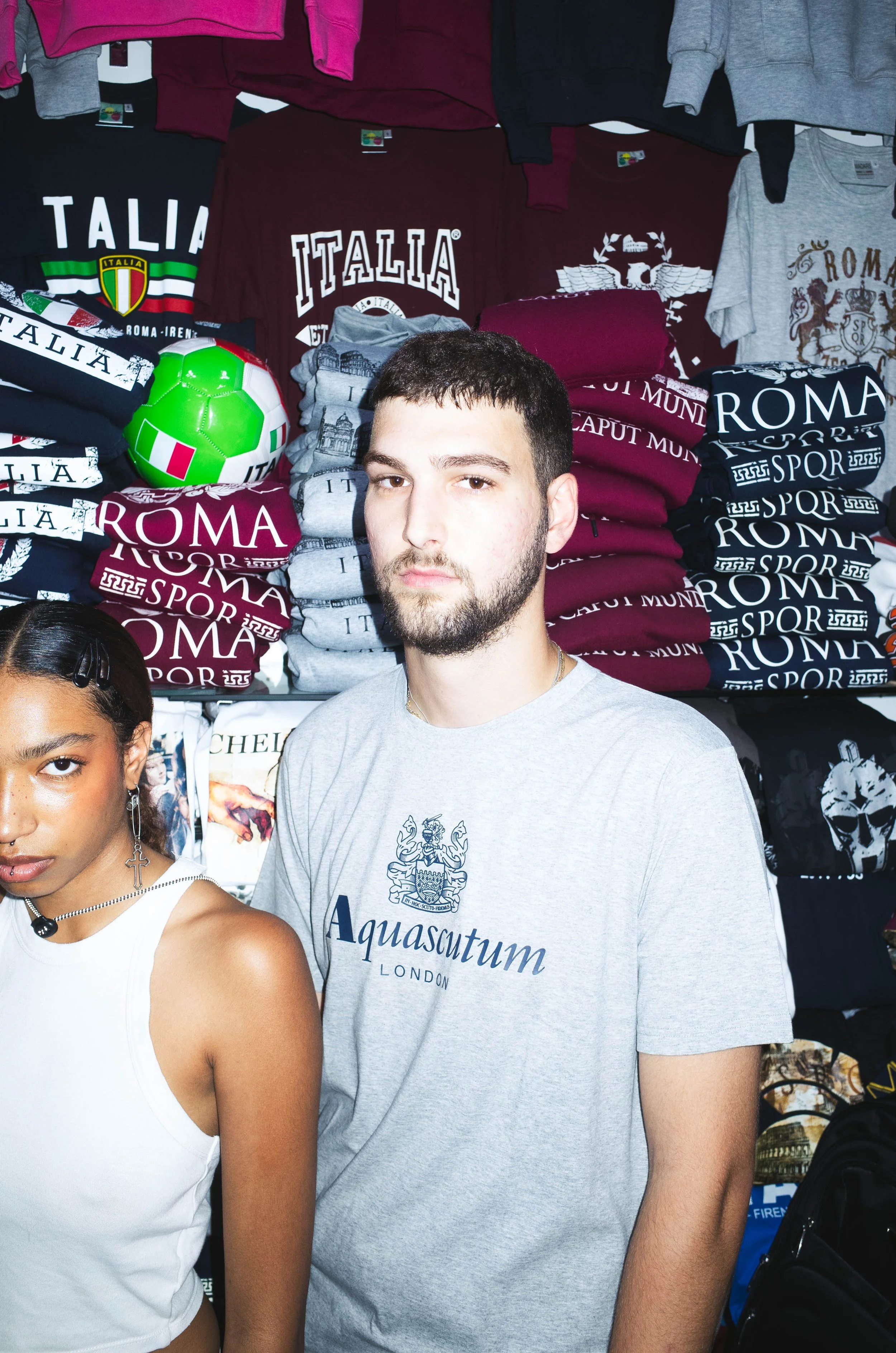 Young man with a beard wearing a gray Aquascutum T-shirt standing in front of stacks of folded Italian-themed clothing and a small green and white soccer ball.