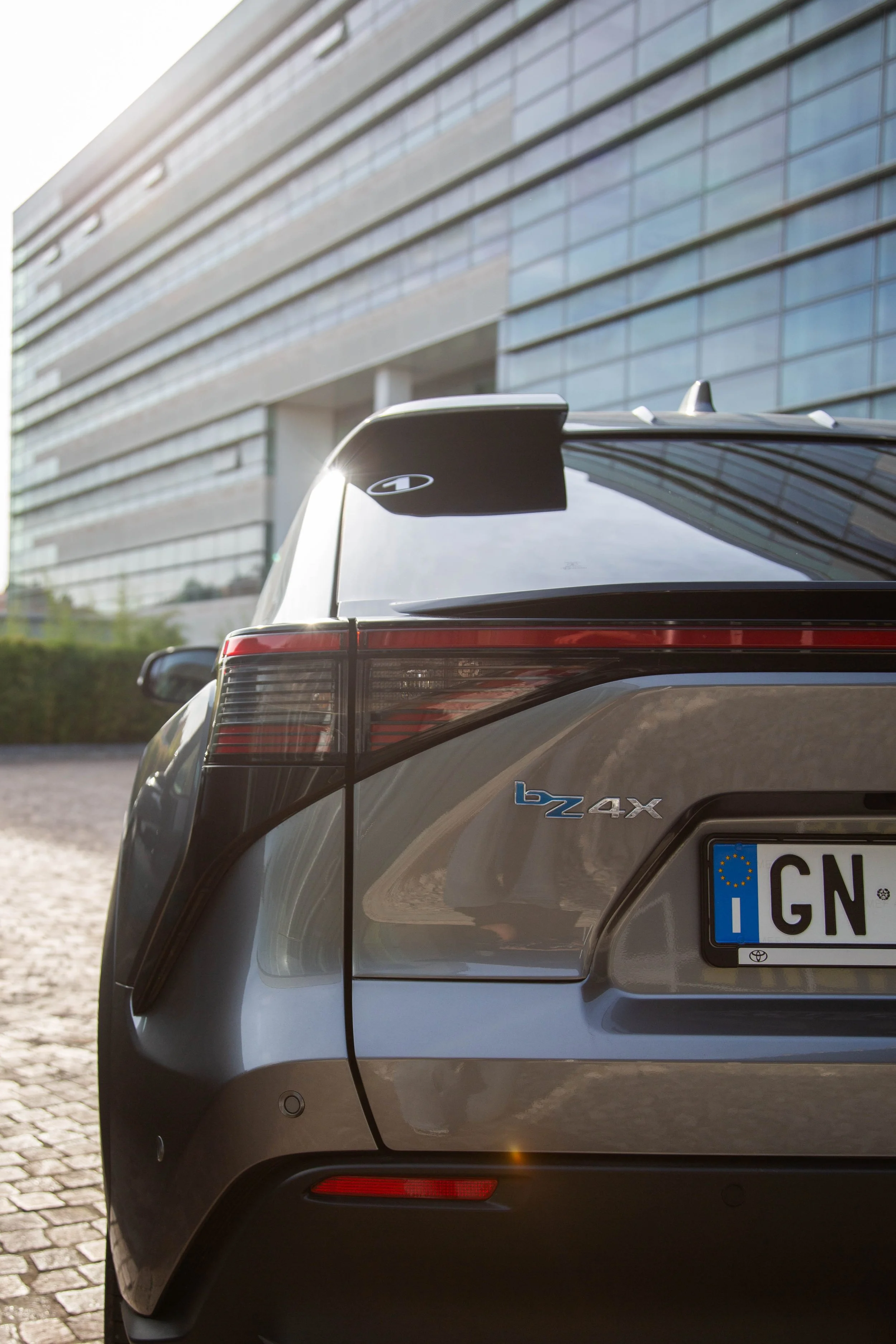 Close-up of the rear of a gray Toyota bZ4X electric vehicle, featuring tail lights, a rear spoiler, and a license plate, parked in front of a modern glass building.