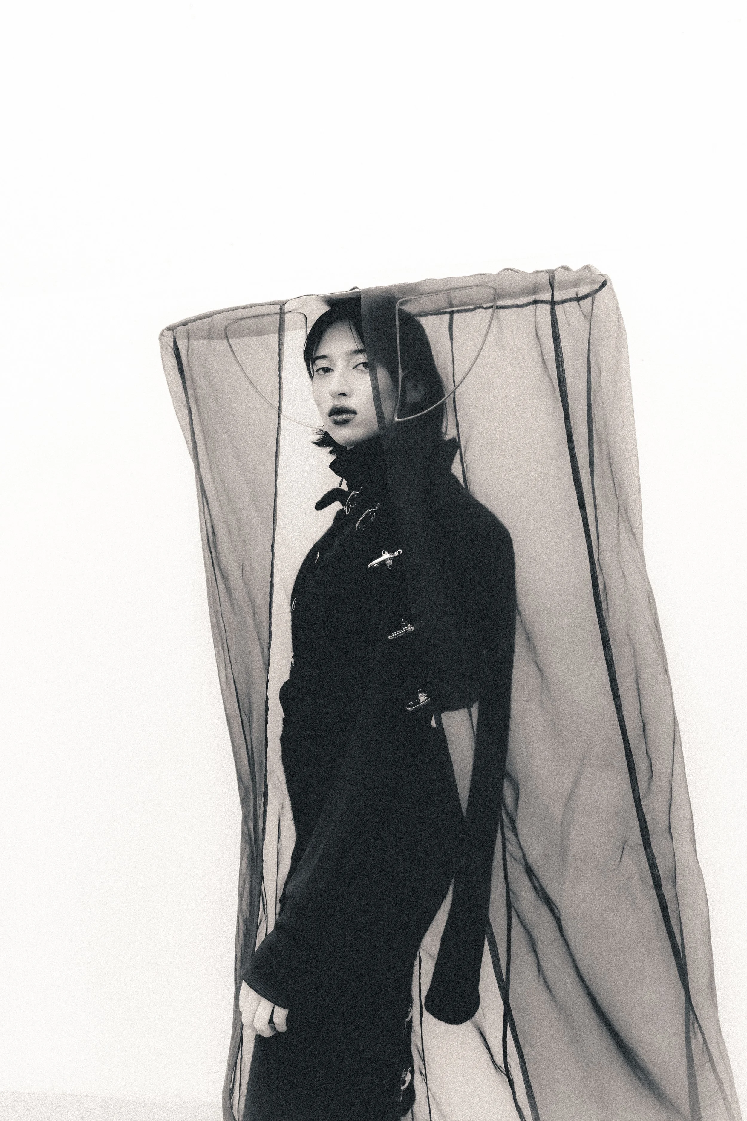 A black-and-white photograph of a woman with dark hair, dressed in a dark outfit with safety pins, standing inside a large, translucent, mesh fabric structure against a plain white background.