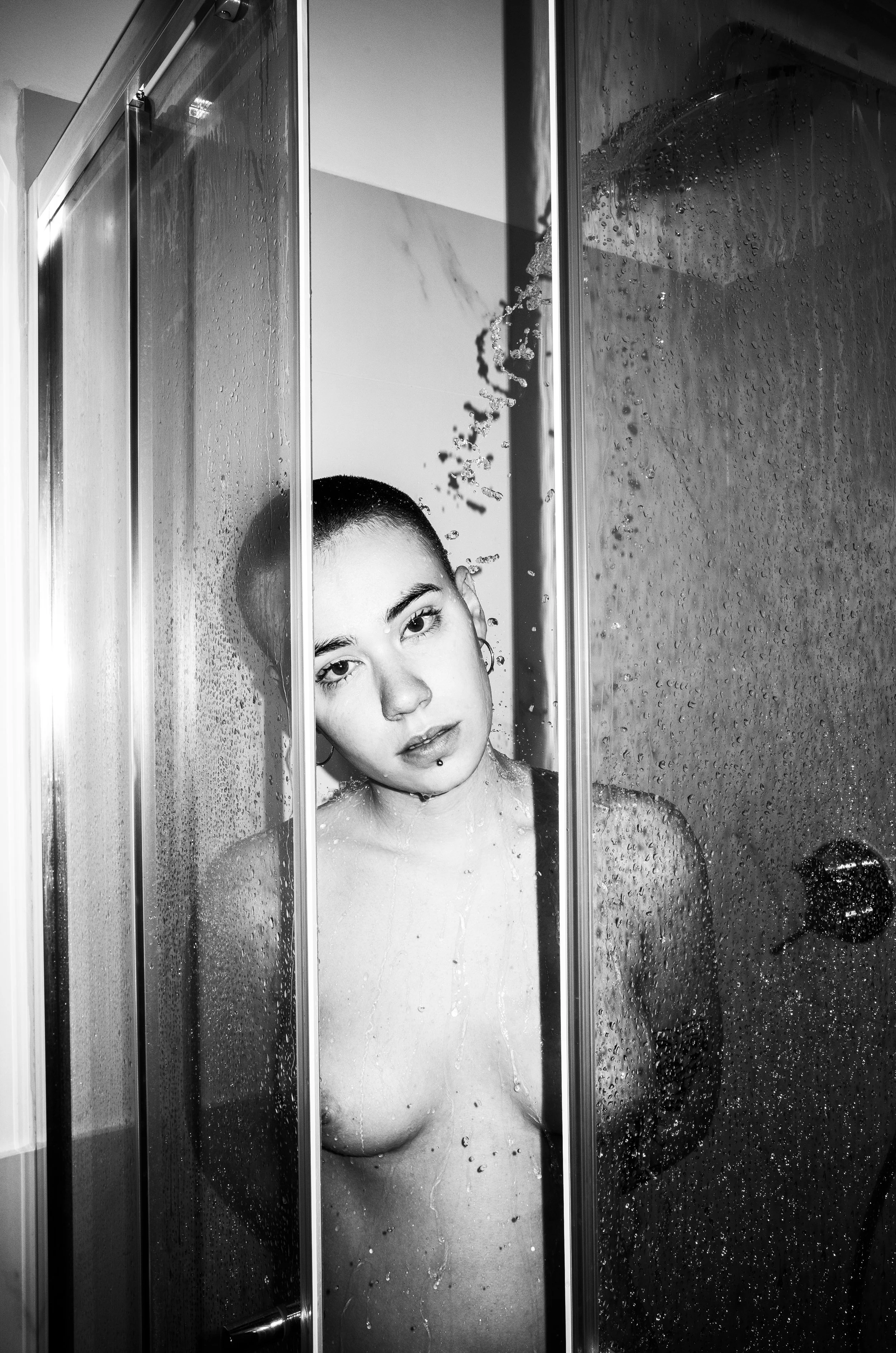 A young woman with short hair and piercings in a shower, looking through the glass door with water droplets.