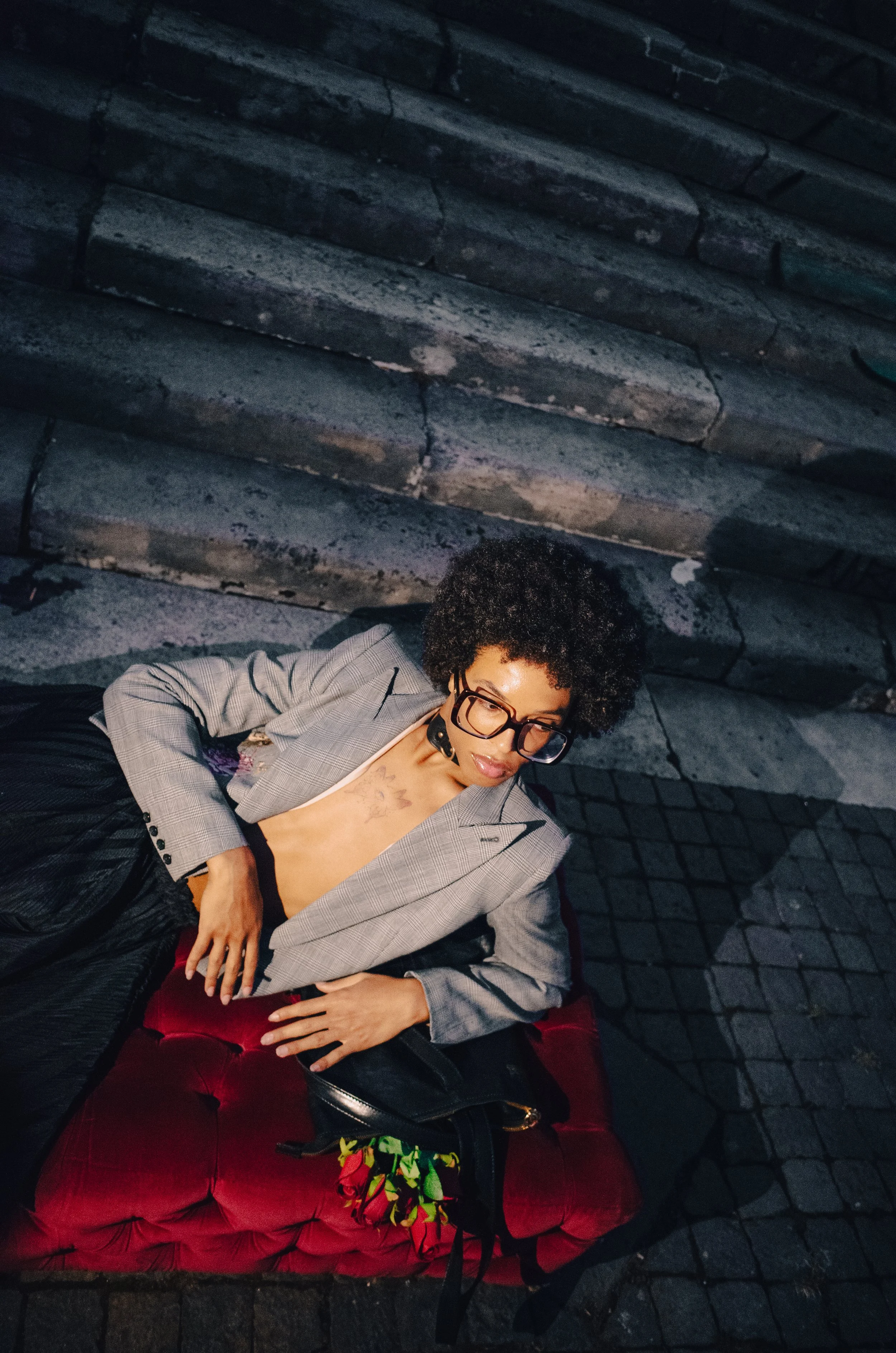 A woman with glasses and curly hair reclining on a red velvet cushion outdoors, wearing a gray blazer, with a tattoo visible on her chest.