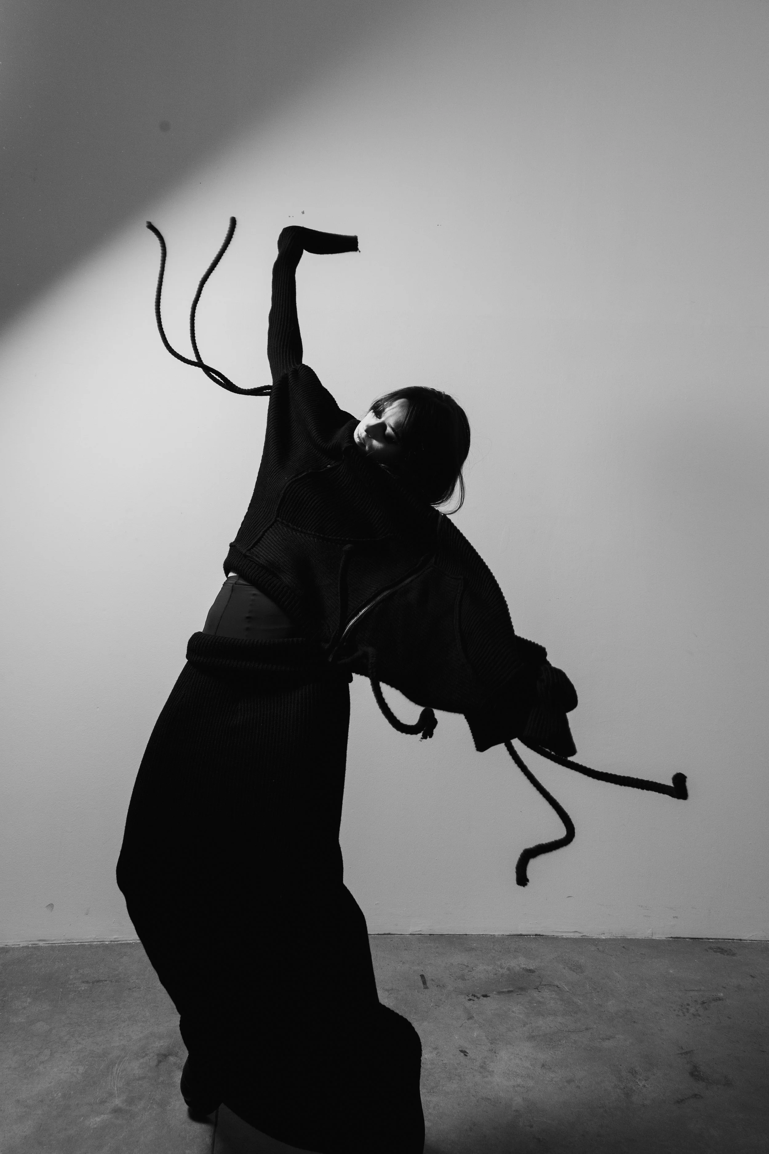 A person dressed in a black outfit, posing dynamically against a plain wall, with some of the clothing's strings hanging loose.