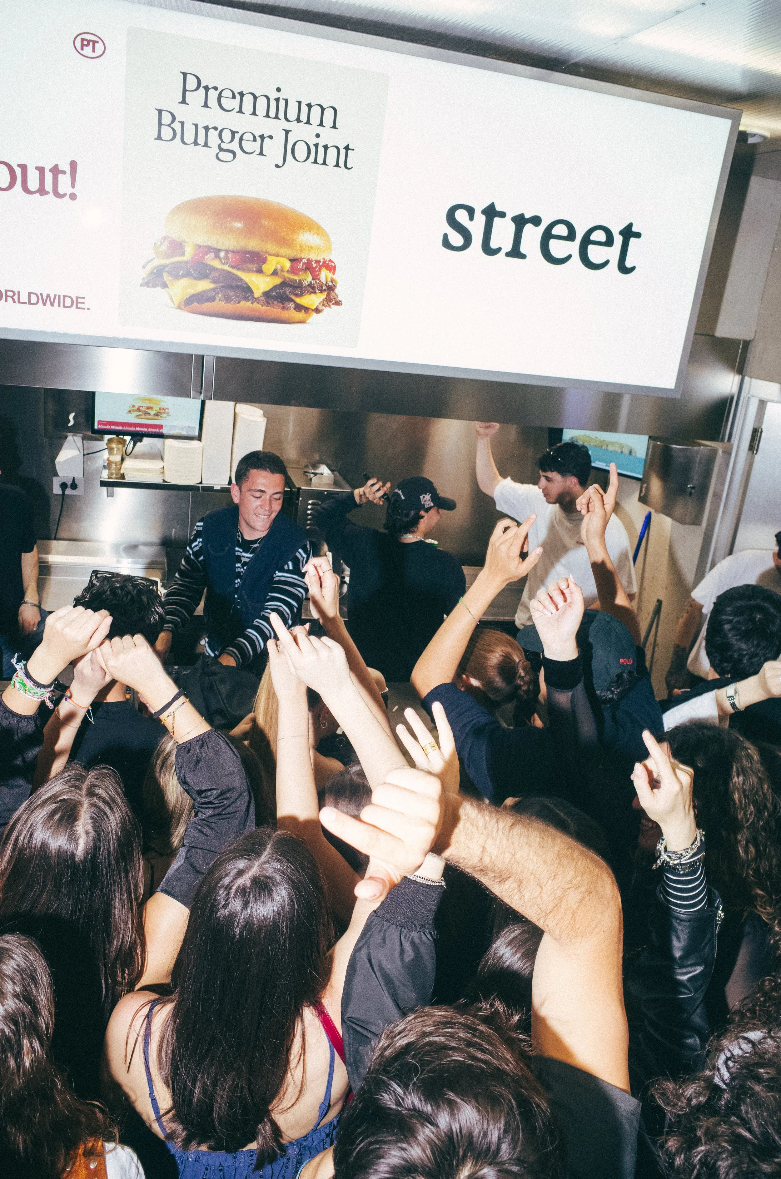 Crowd of excited people at a fast-food restaurant with a sign reading 'Premium Burger Joint' and 'street'.