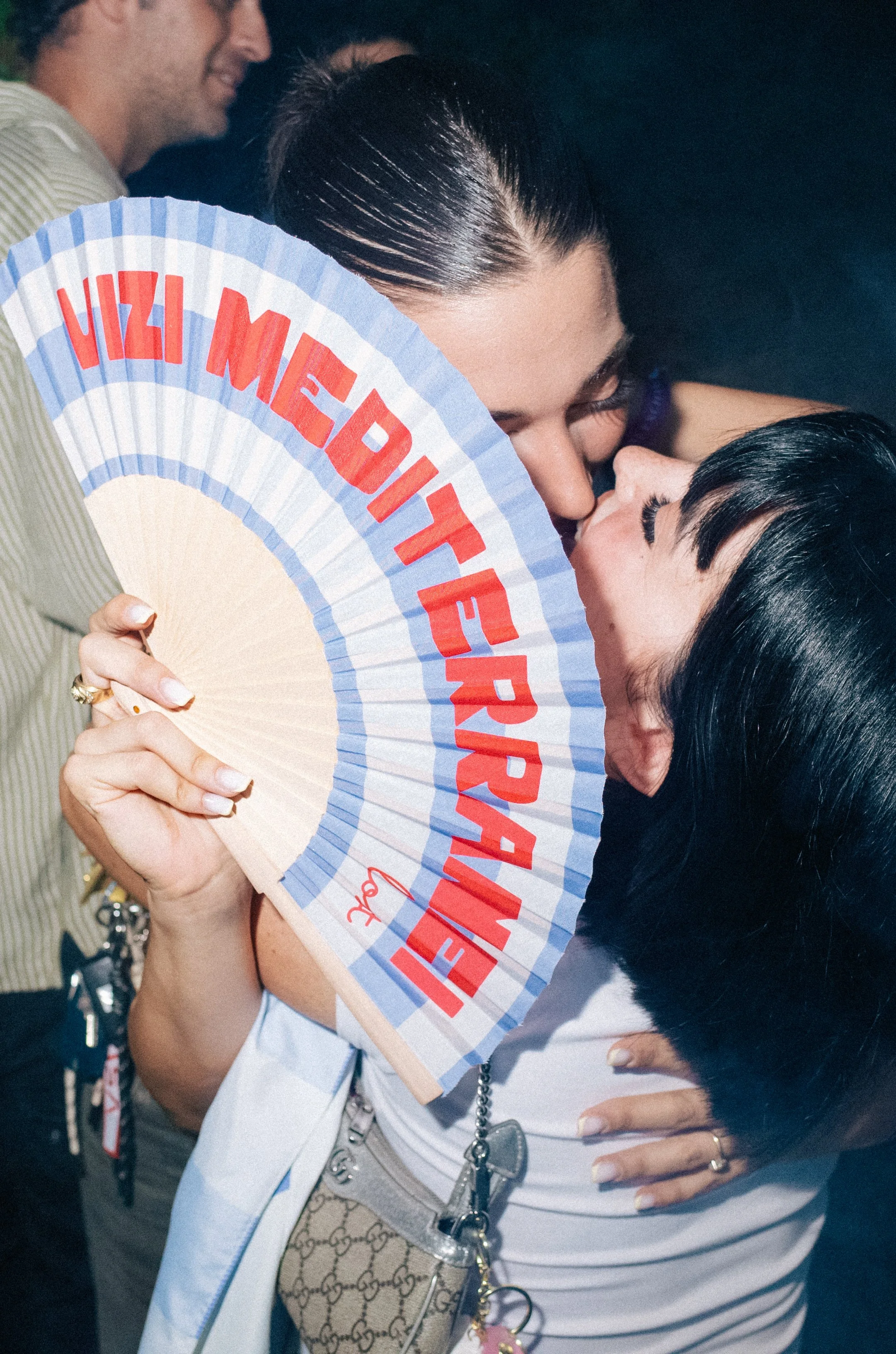 Two women kissing, one holding a fan with the phrase 'HIZI MEDIA' written on it, in a close and intimate moment.