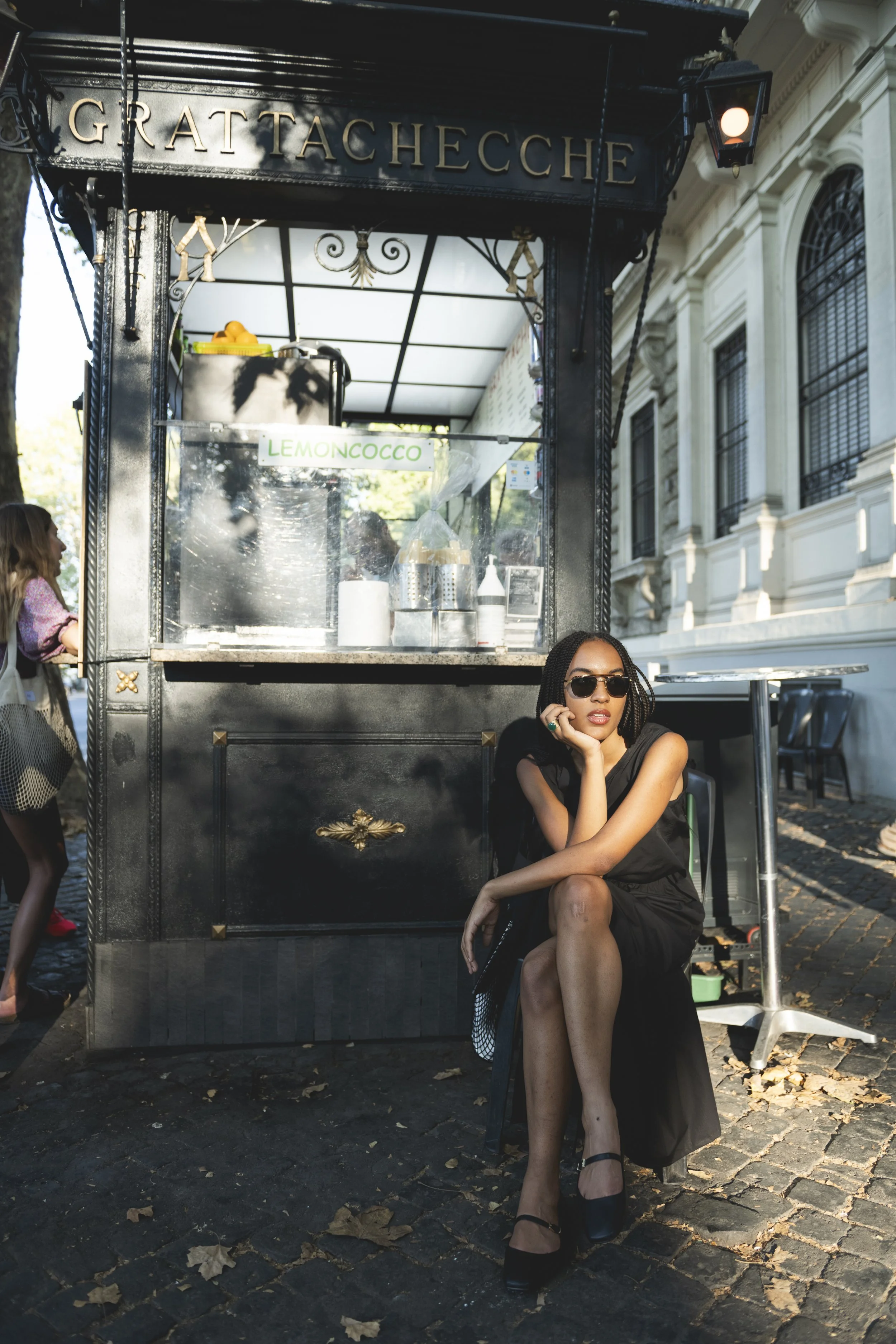 A woman in black dress and sunglasses sitting on a chair next to a street food cart, with a building and another person in the background.