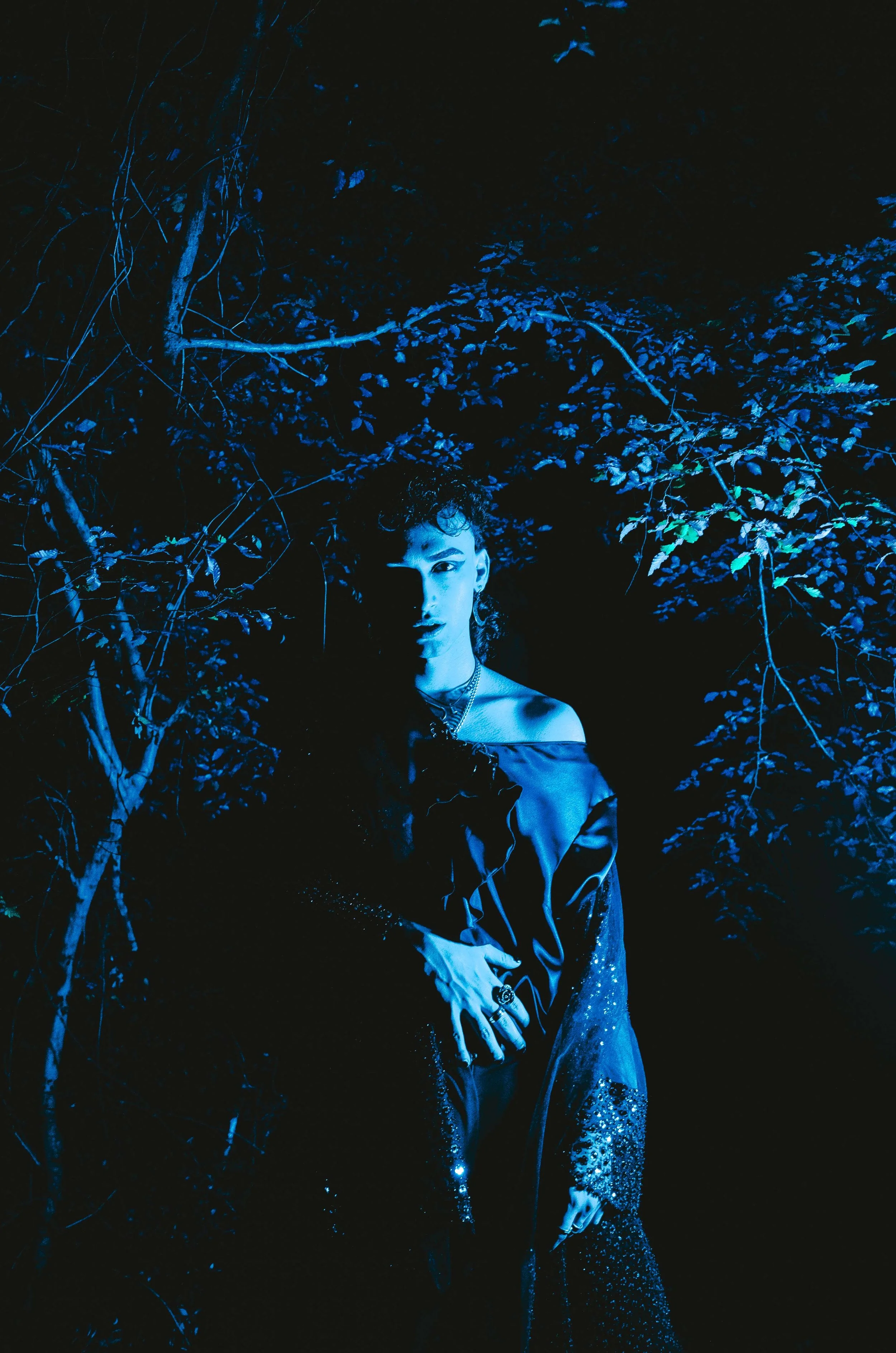 A young man standing in a dark forest, illuminated with blue lighting, wearing a dark, shiny, off-shoulder top and jewelry, surrounded by trees and foliage.