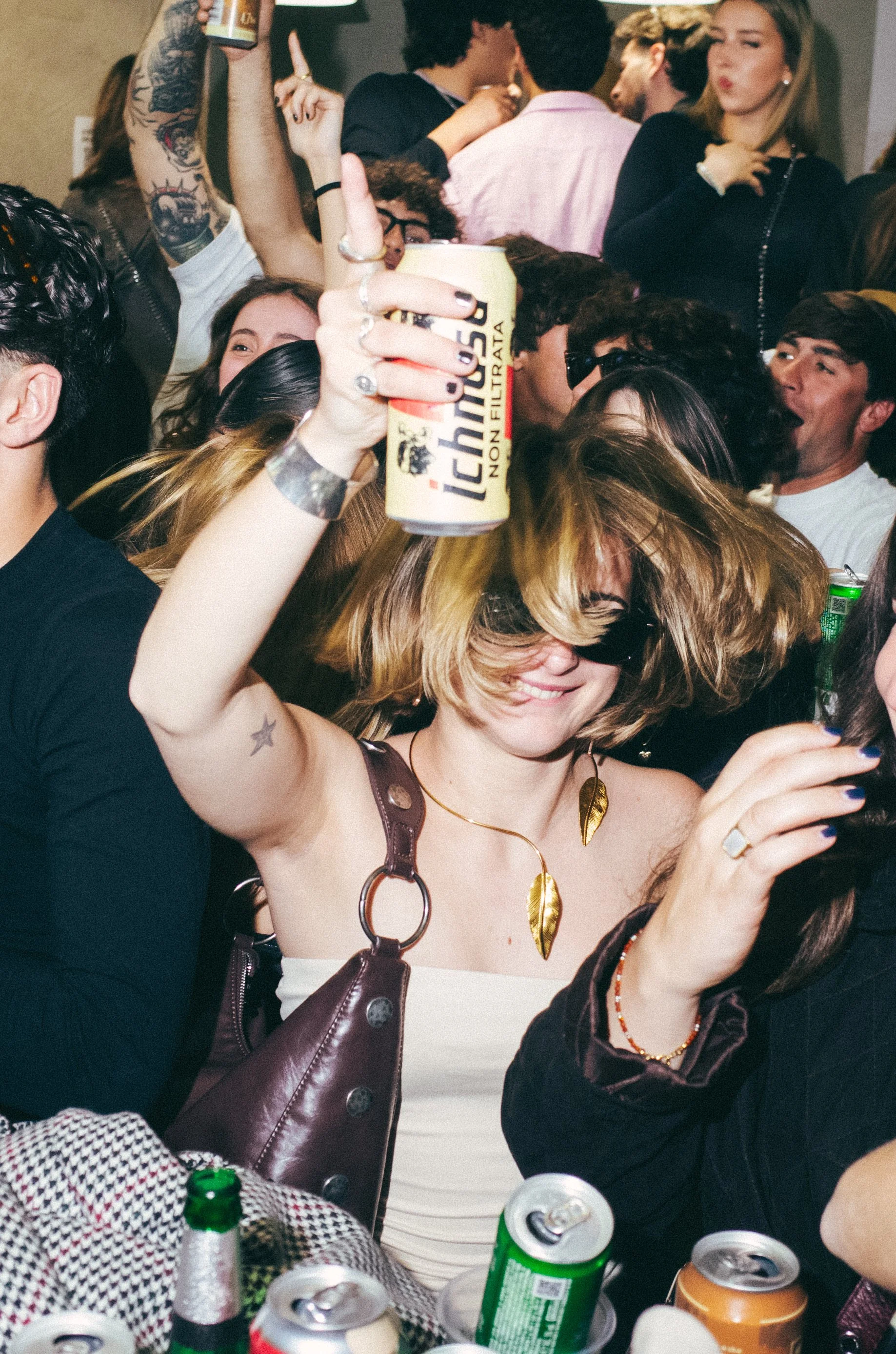 A woman at a crowded party dancing, smiling, and holding a can of Tecate beer in the air. She has light brown hair, sunglasses, and is wearing a white top with gold leaf earrings and a necklace with large gold leaves.