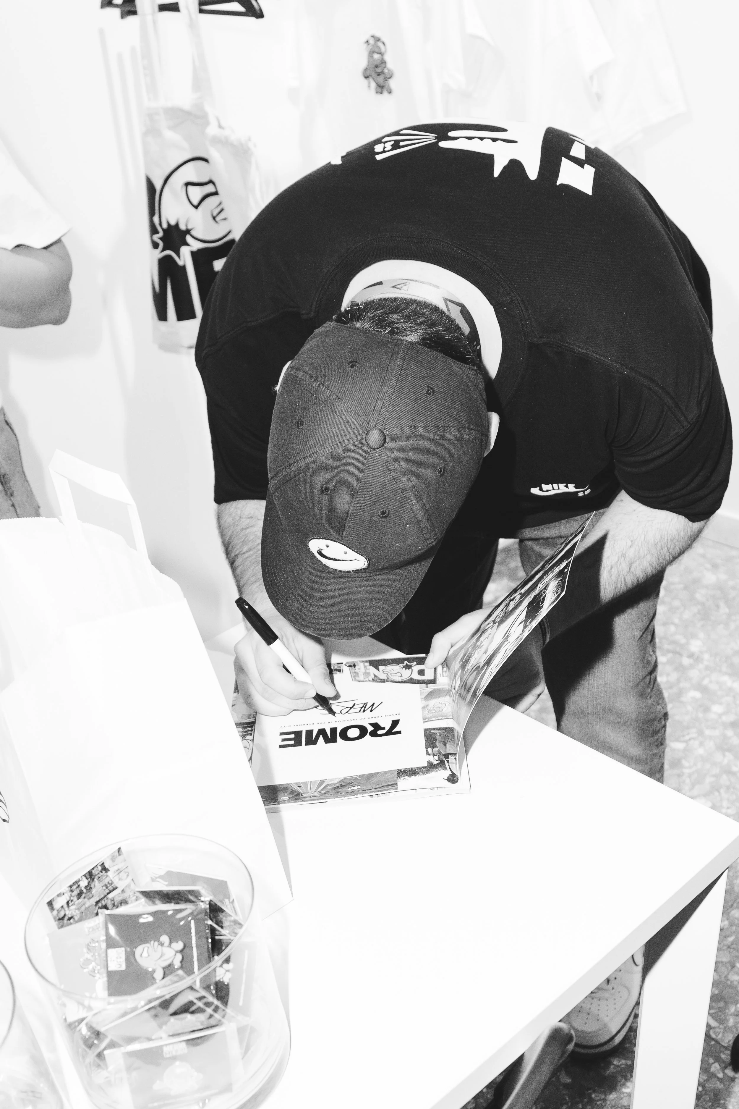 Man wearing a cap signing a poster at a table, with a sign that says 'ROWDIE' and a bowl containing various items next to him.