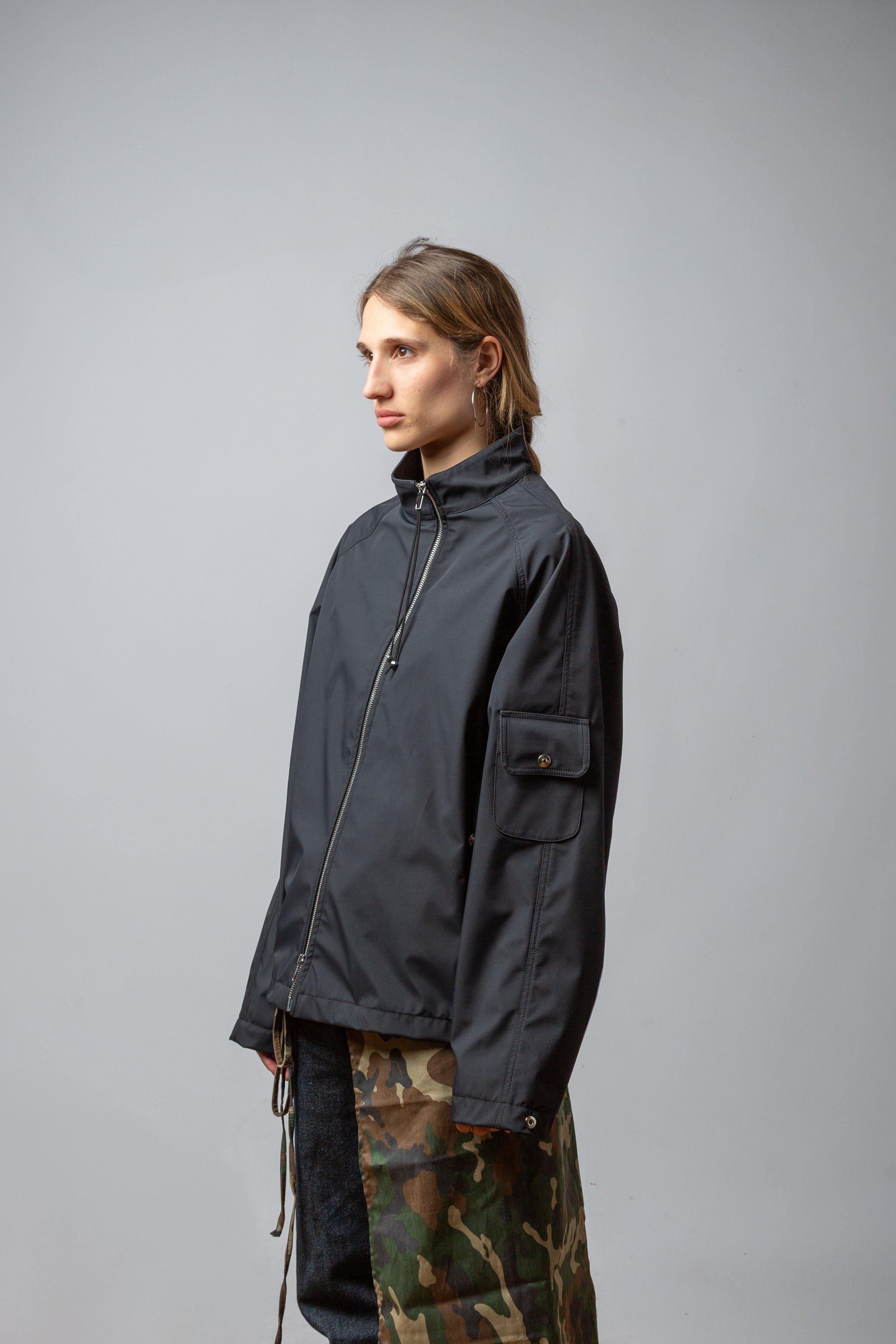 A woman with light brown hair and hoop earrings, wearing a black zip-up jacket and camouflage pants, standing against a plain grey background.