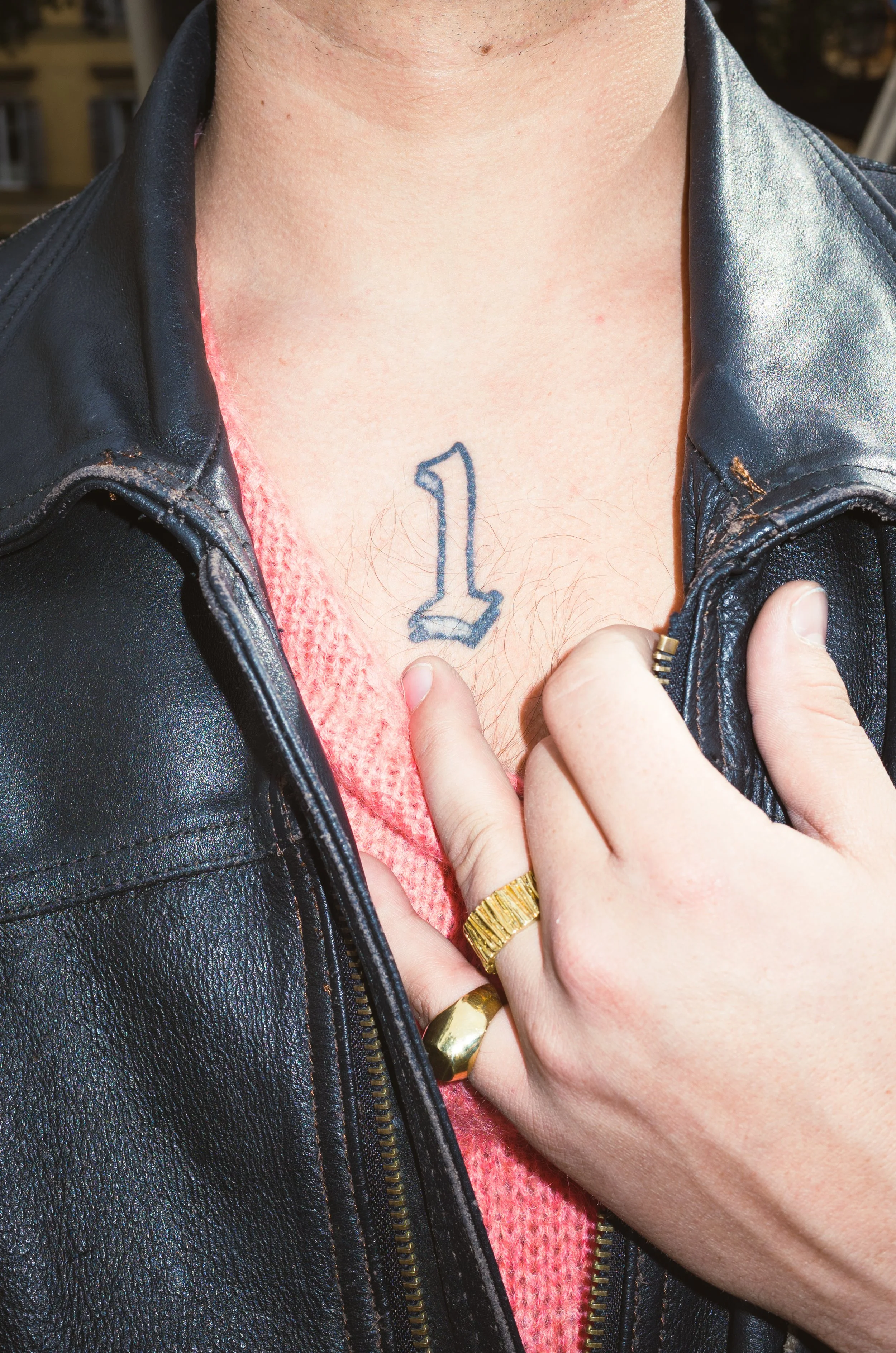 Close-up of a person's chest showcasing a tattoo of the number 1 on the sternum area, partially covered by their hand pulling a black leather jacket open, revealing a red knitted sweater underneath.