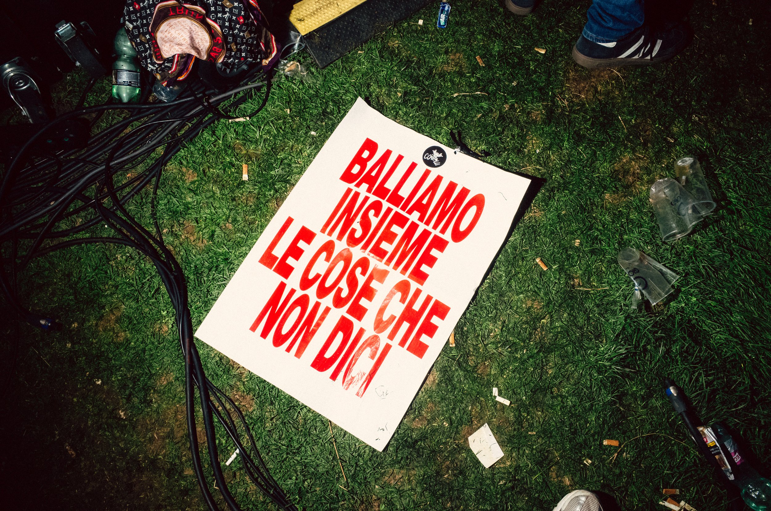 A protest sign on the ground with Italian text in bold red letters, surrounded by scattered trash and waste on grass, with cables, bottles, and shoes nearby.