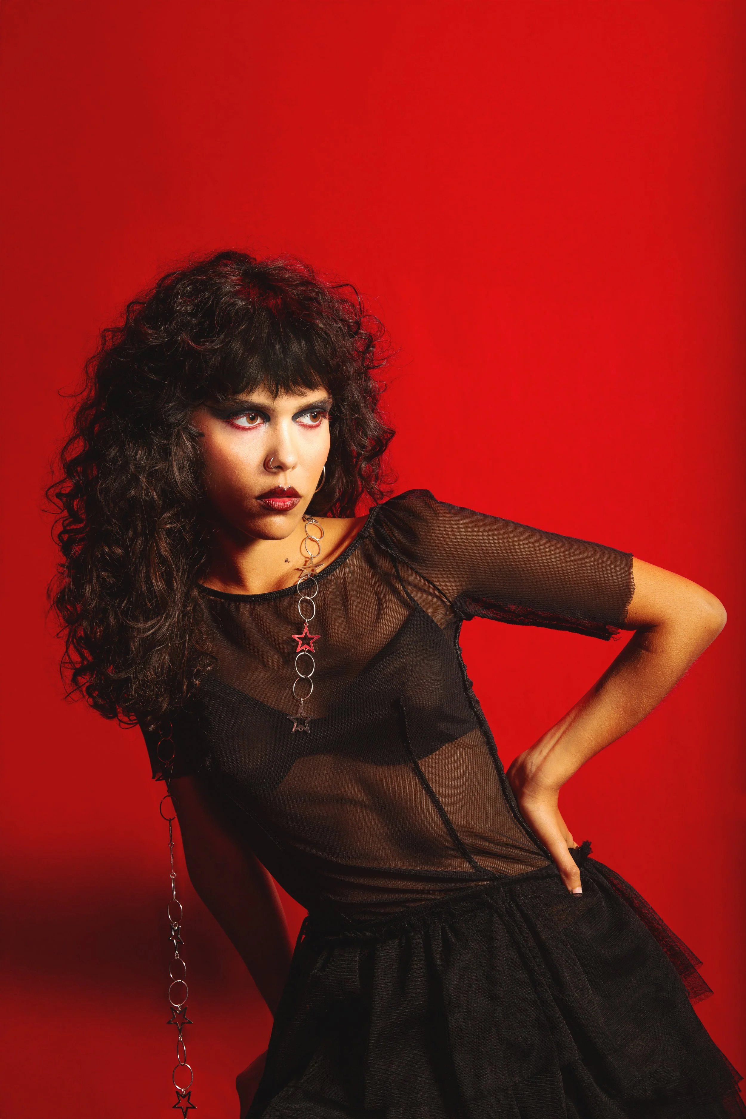 Young woman with curly dark hair wearing a sheer black top, black tulle skirt, and star jewelry against red background.