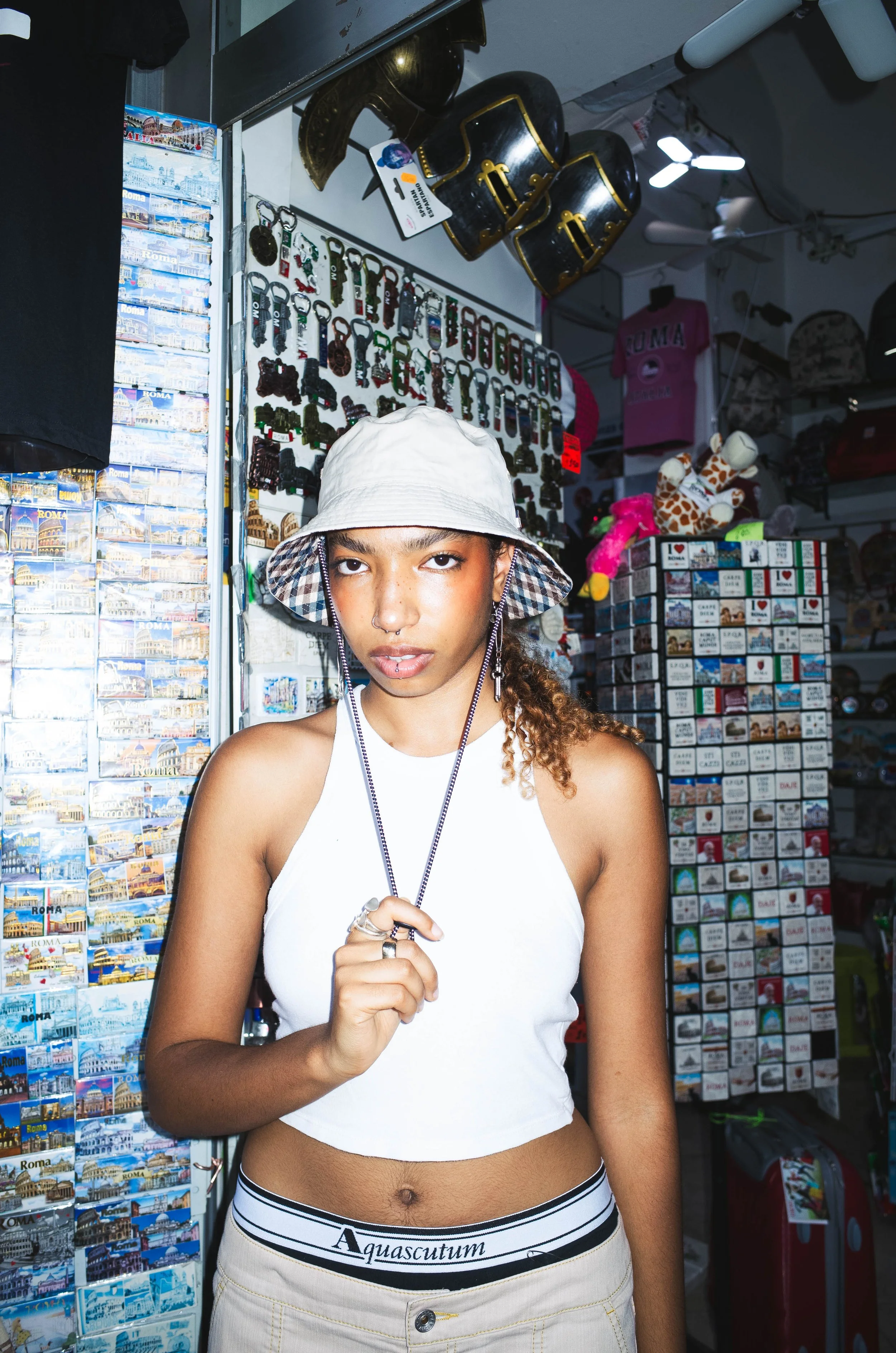 A young woman with curly hair, wearing a beige bucket hat, white crop top, beige pants with a black and white waistband, and jewelry, standing inside a gift shop surrounded by souvenirs, keychains, and stuffed animals.