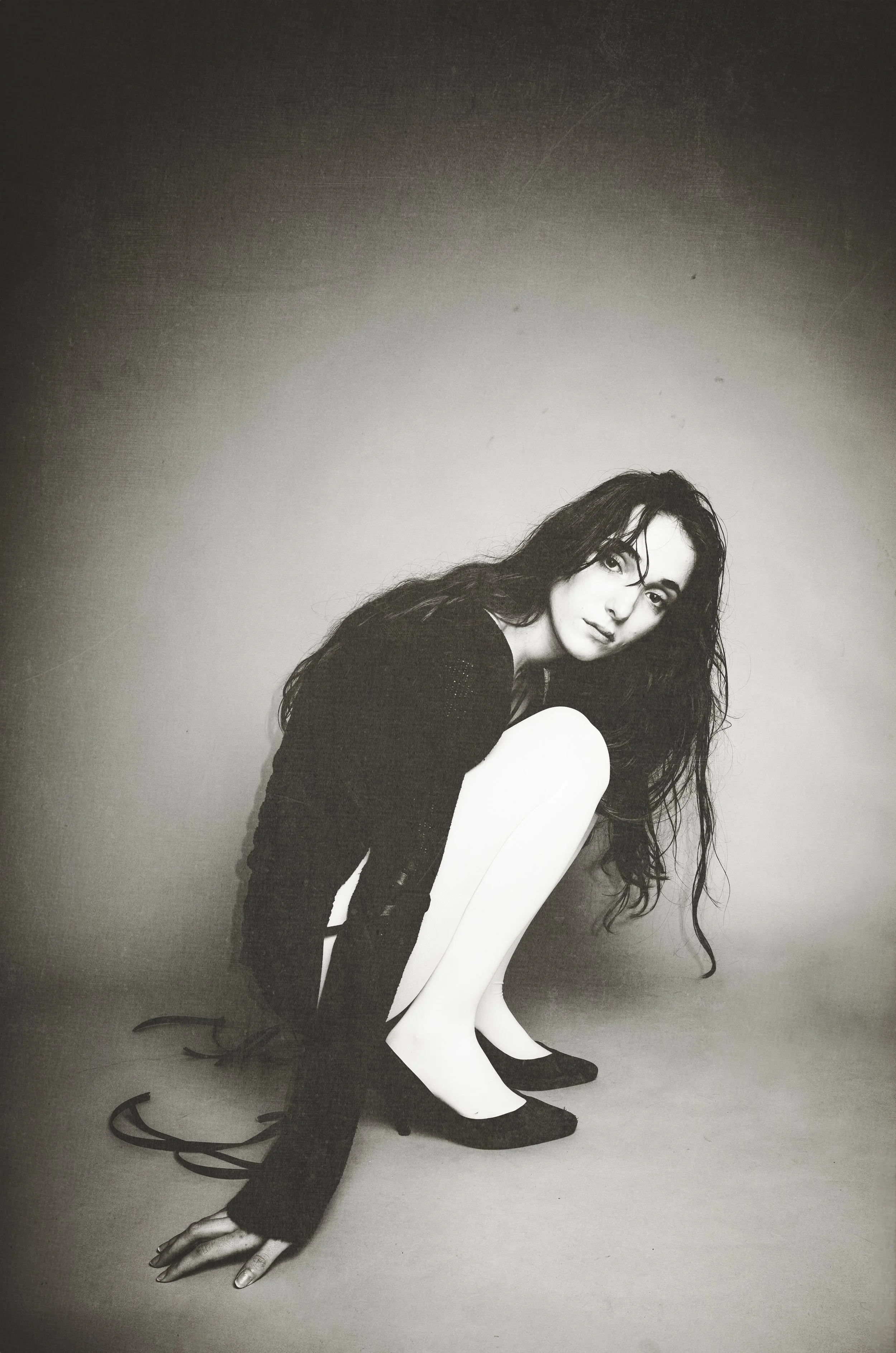 Black and white photograph of a woman with long curly hair, crouching with one hand on the ground and her knees drawn up, dressed in black shoes, a black top, and white pants, against a simple gradient background.