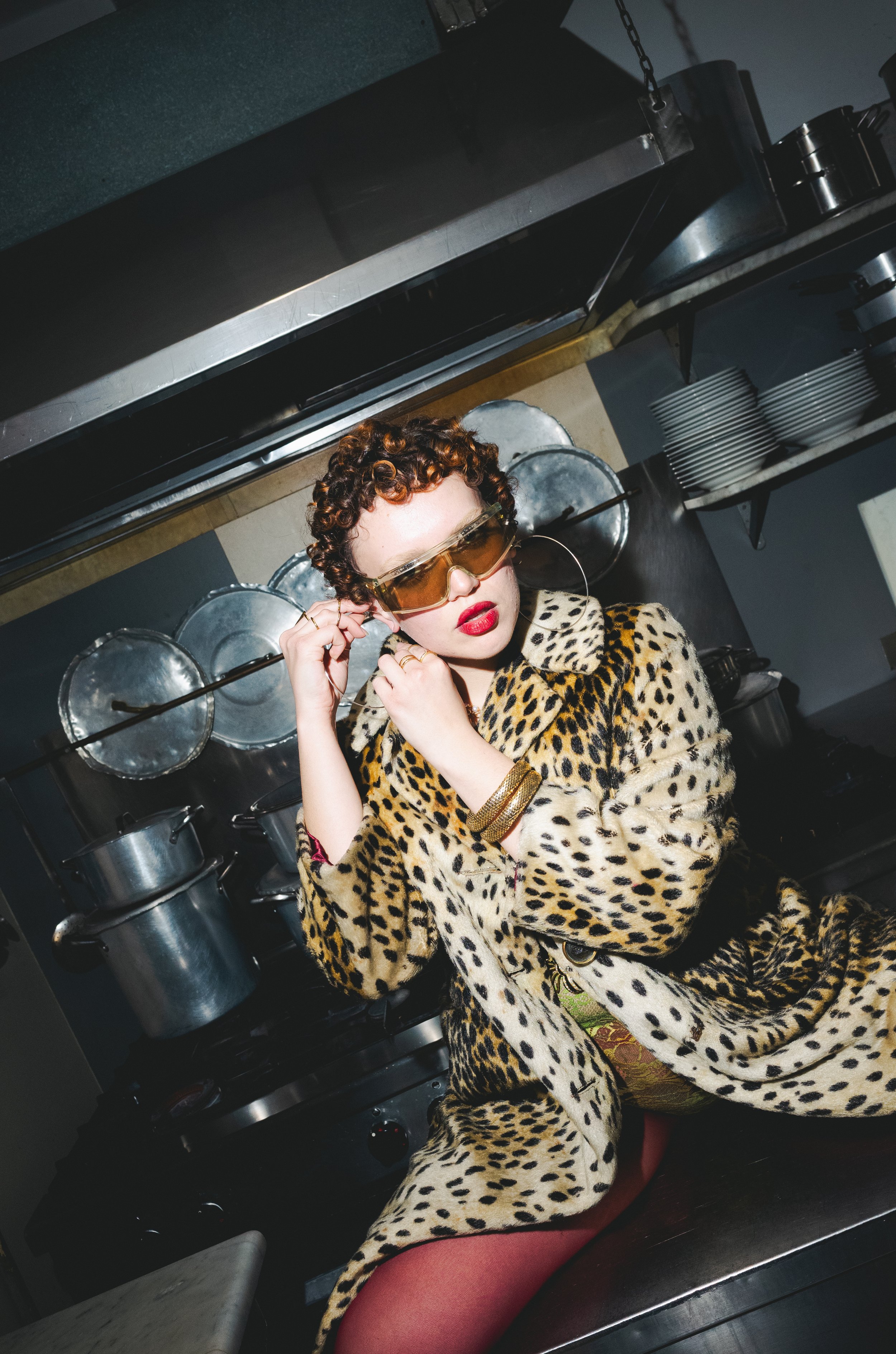 Woman with curly hair wearing oversized sunglasses and a leopard print coat, sitting in a kitchen with pots and pans in the background.