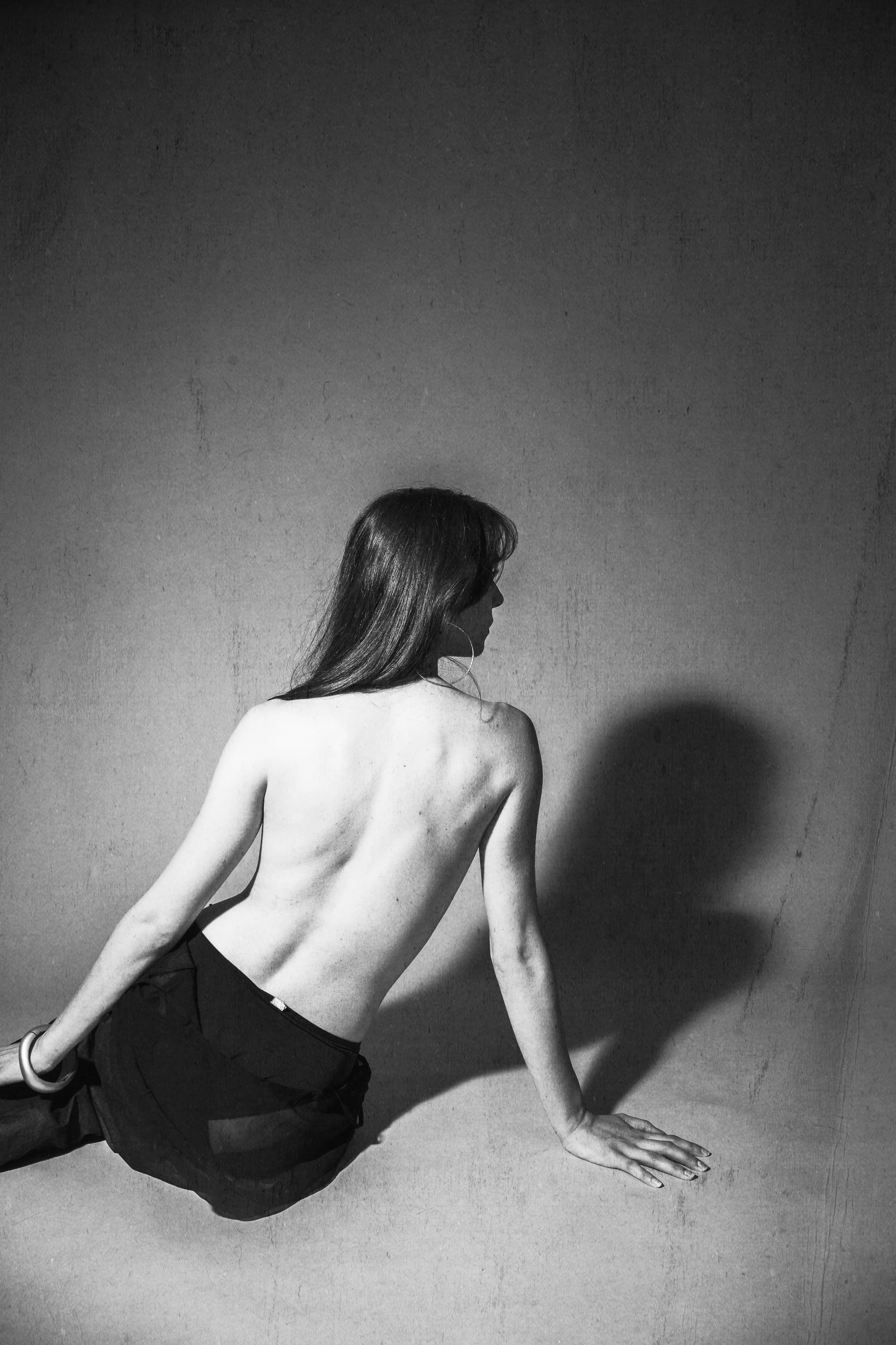 A woman with long hair, topless and wearing dark pants, is sitting on the ground with one hand on the floor and her back facing the camera, casting a shadow on the wall.