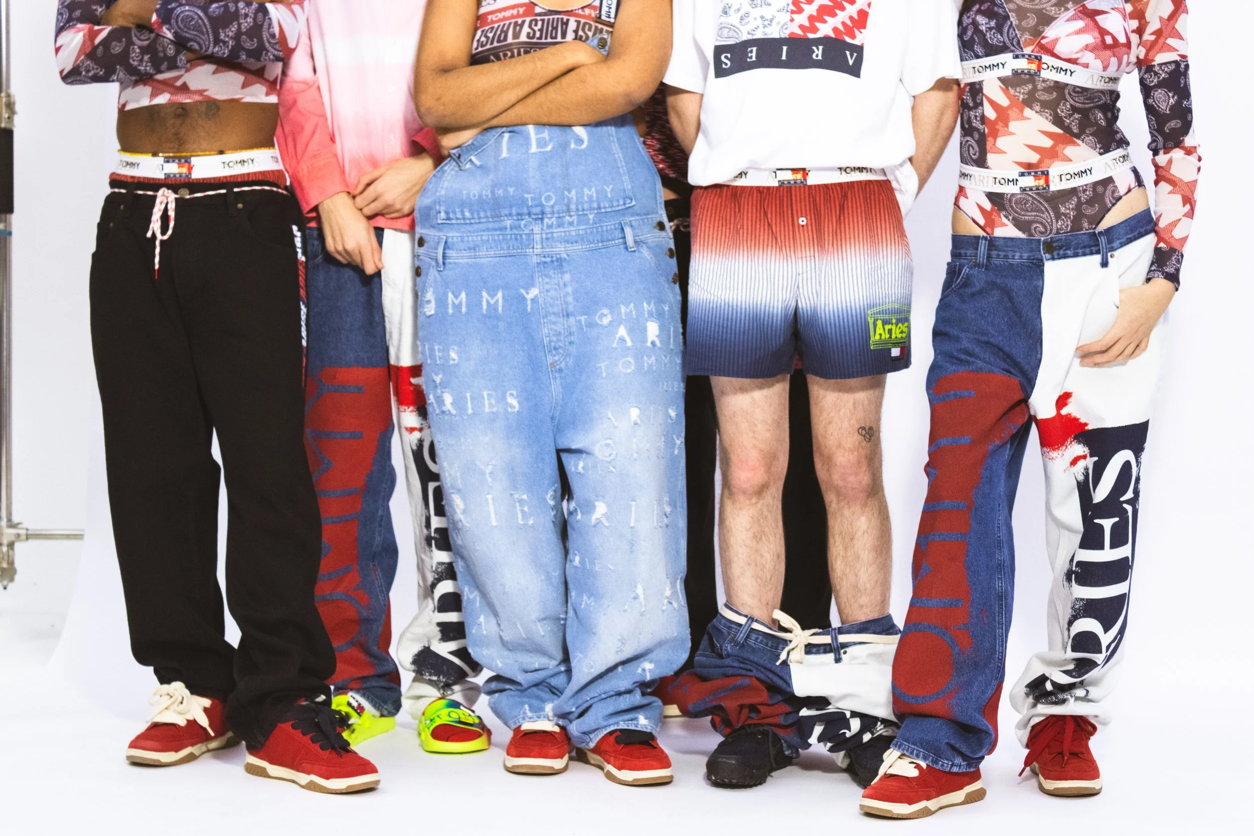 Close-up of five people showing their colorful, branded fashion jeans and sneakers, with their torsos and legs visible against a white background.